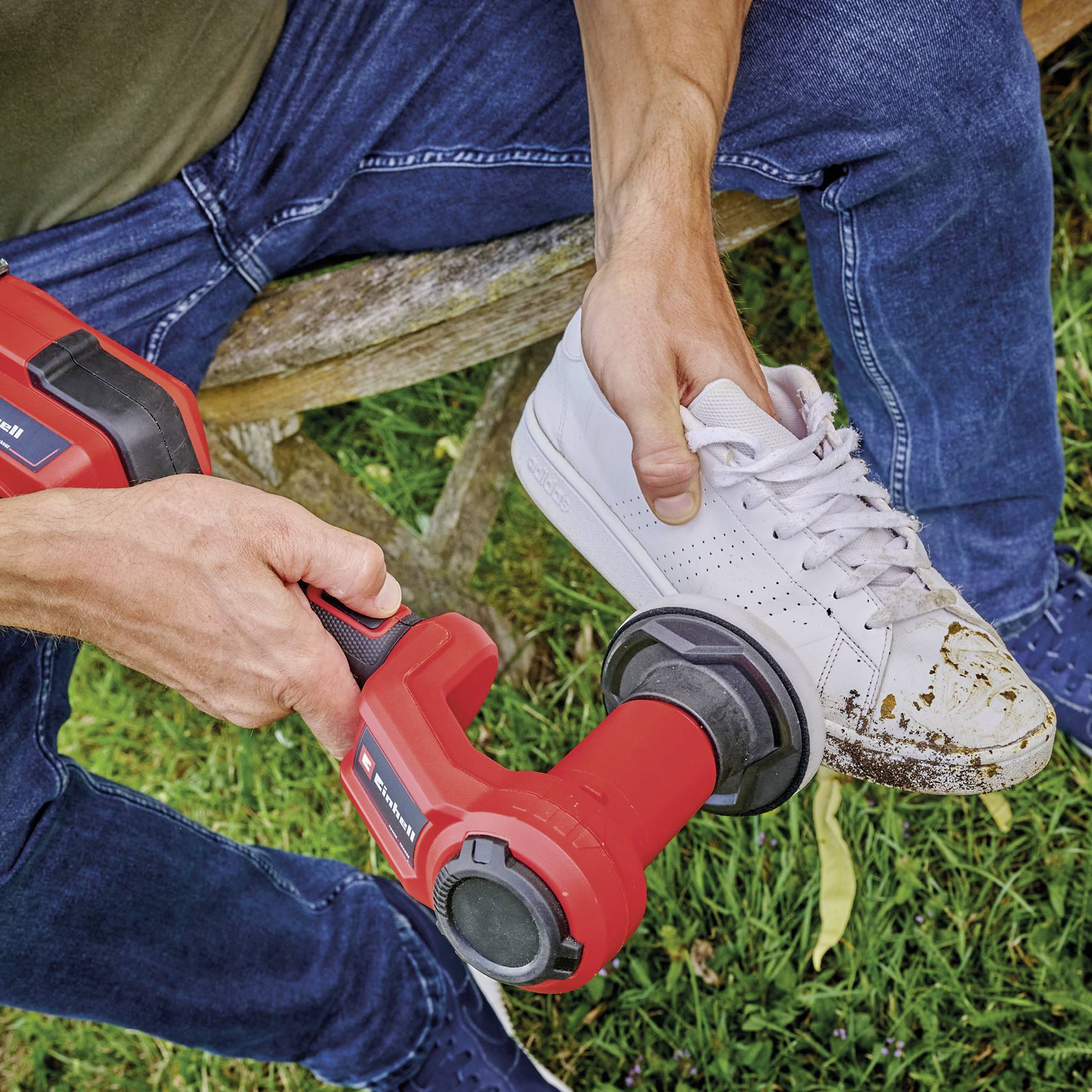 Une personne nettoie une chaussure de sport blanche avec un appareil de nettoyage électrique rouge sur une pelouse.