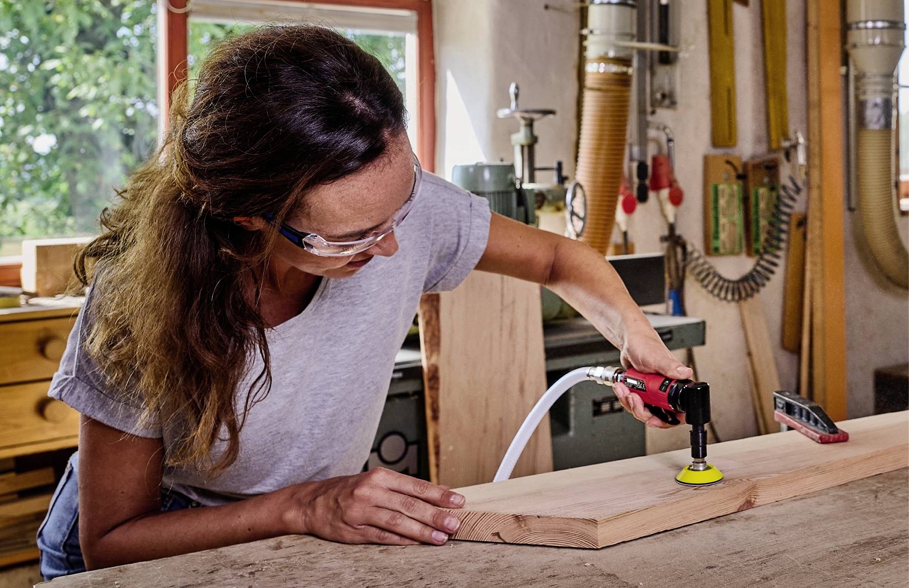 Une femme porte des lunettes de protection et ponce une planche en bois à l'aide d'une ponceuse dans un atelier.