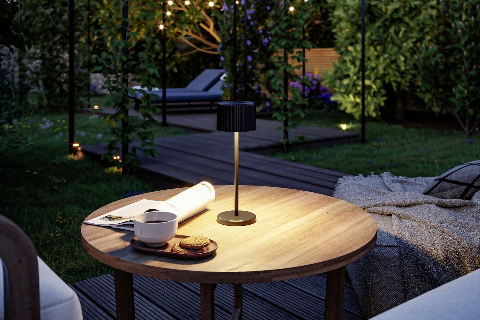 Une table ronde en bois dans le jardin avec une tasse de café, des biscuits, une lampe et un livre. Des plantes et un transat sont visibles en arrière-plan.