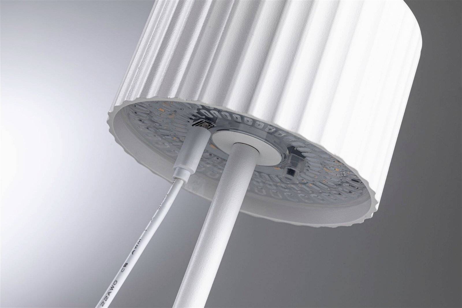 Lampe de table blanche avec un abat-jour strié, vue en contre-plongée. Un câble descend vers la base de la lampe. Arrière-plan en nuances de gris.
