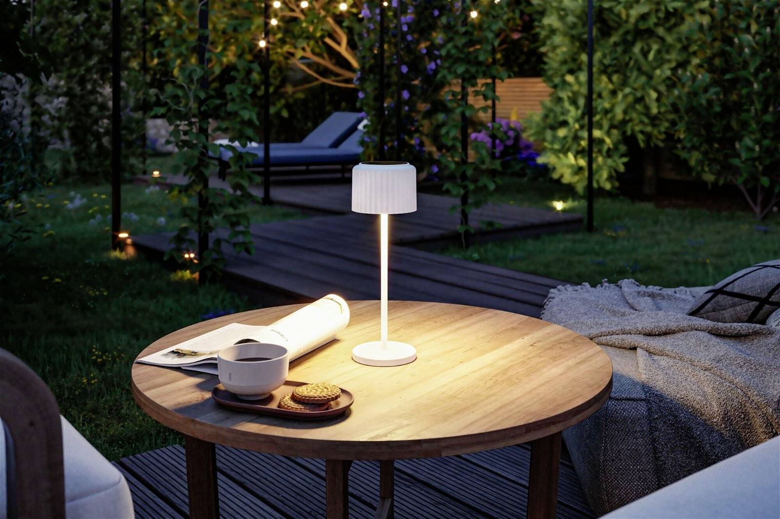 Une table ronde en bois dans la lumière du soir au jardin, avec une petite lampe, une tasse de café, des biscuits et un livre ouvert. À l'arrière-plan, une terrasse en bois et des plantes.