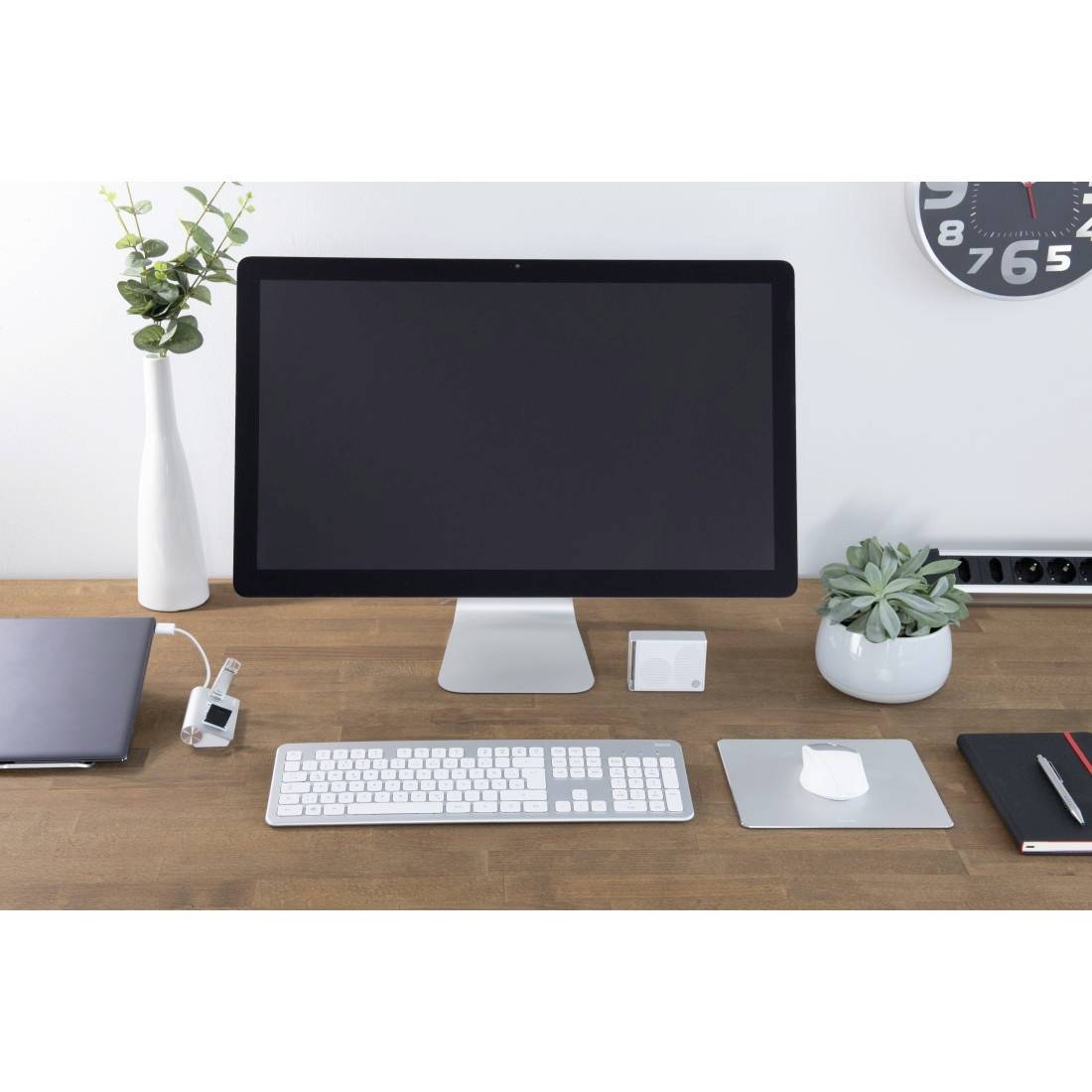Un bureau bien rangé avec un grand écran, un clavier, une souris et des plantes. L'horloge murale indique peu après neuf heures.