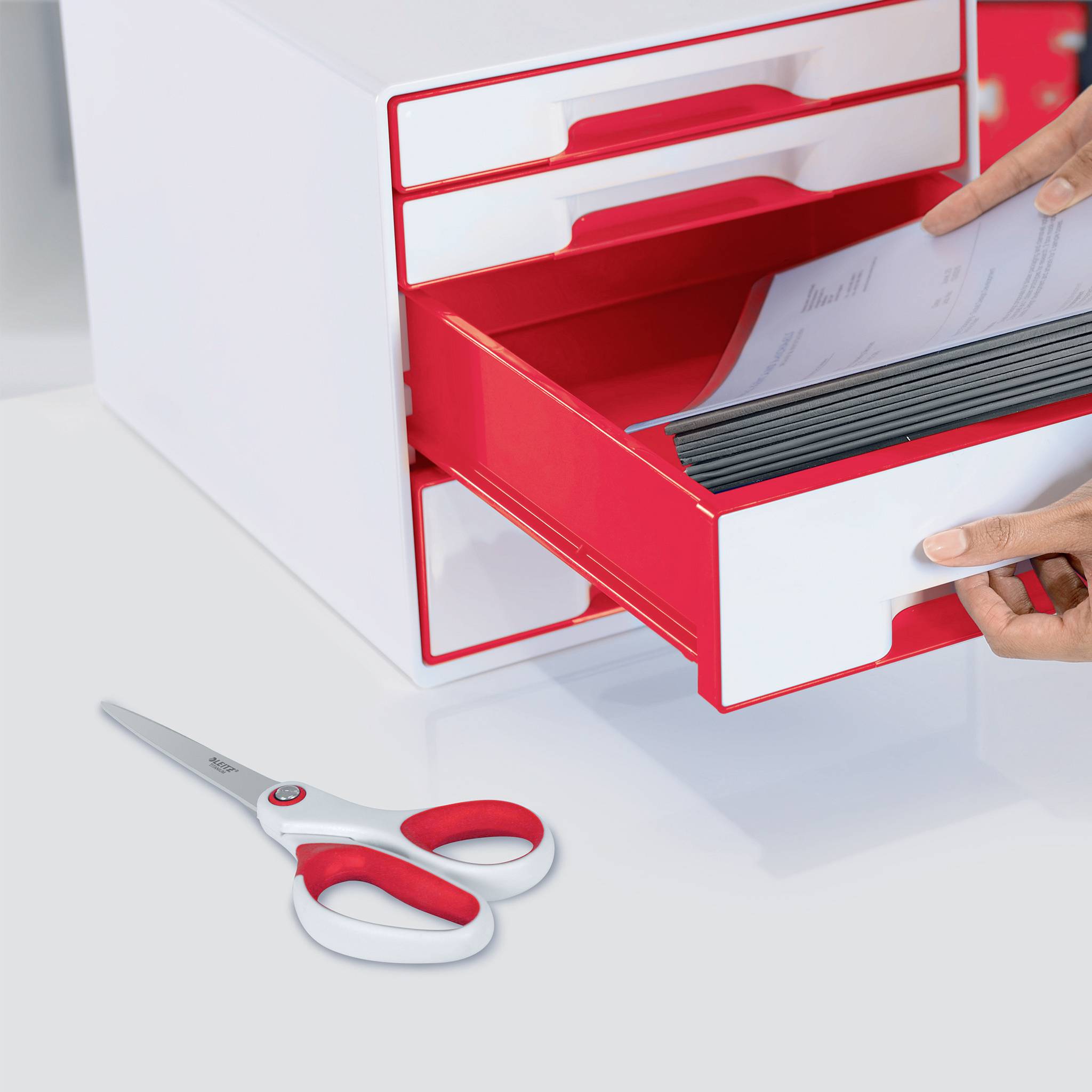 Organisateur de tiroir rouge avec un compartiment ouvert rempli de dossiers, à côté une paire de ciseaux avec une poignée rouge, sur un fond blanc.