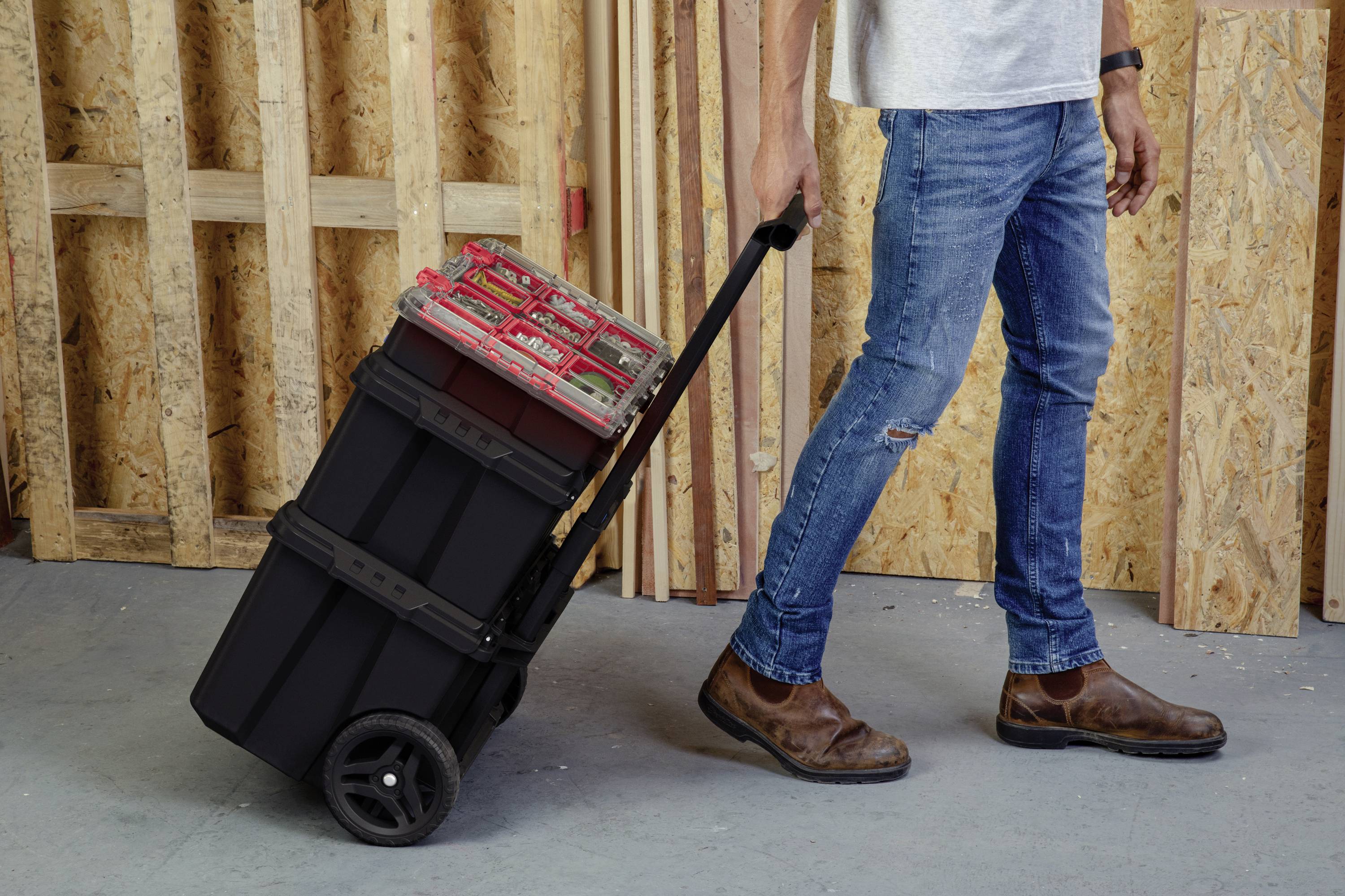 Une personne pousse un chariot à outils noir avec de grandes roues sur un sol en béton. Des planches de bois sont visibles en arrière-plan.