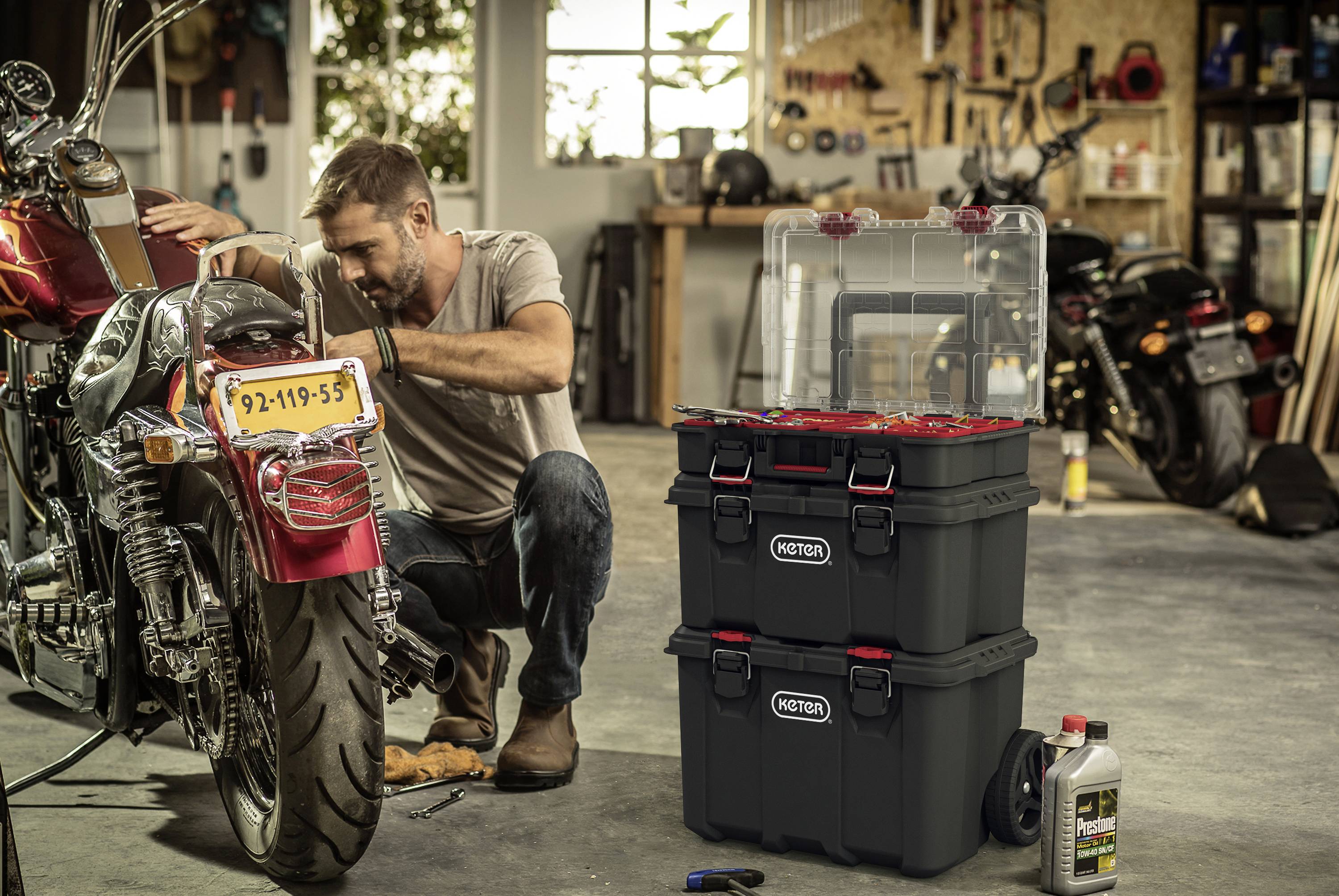 Un homme répare une moto dans un atelier. À côté de lui se trouve une boîte à outils ouverte et une bouteille d'huile.