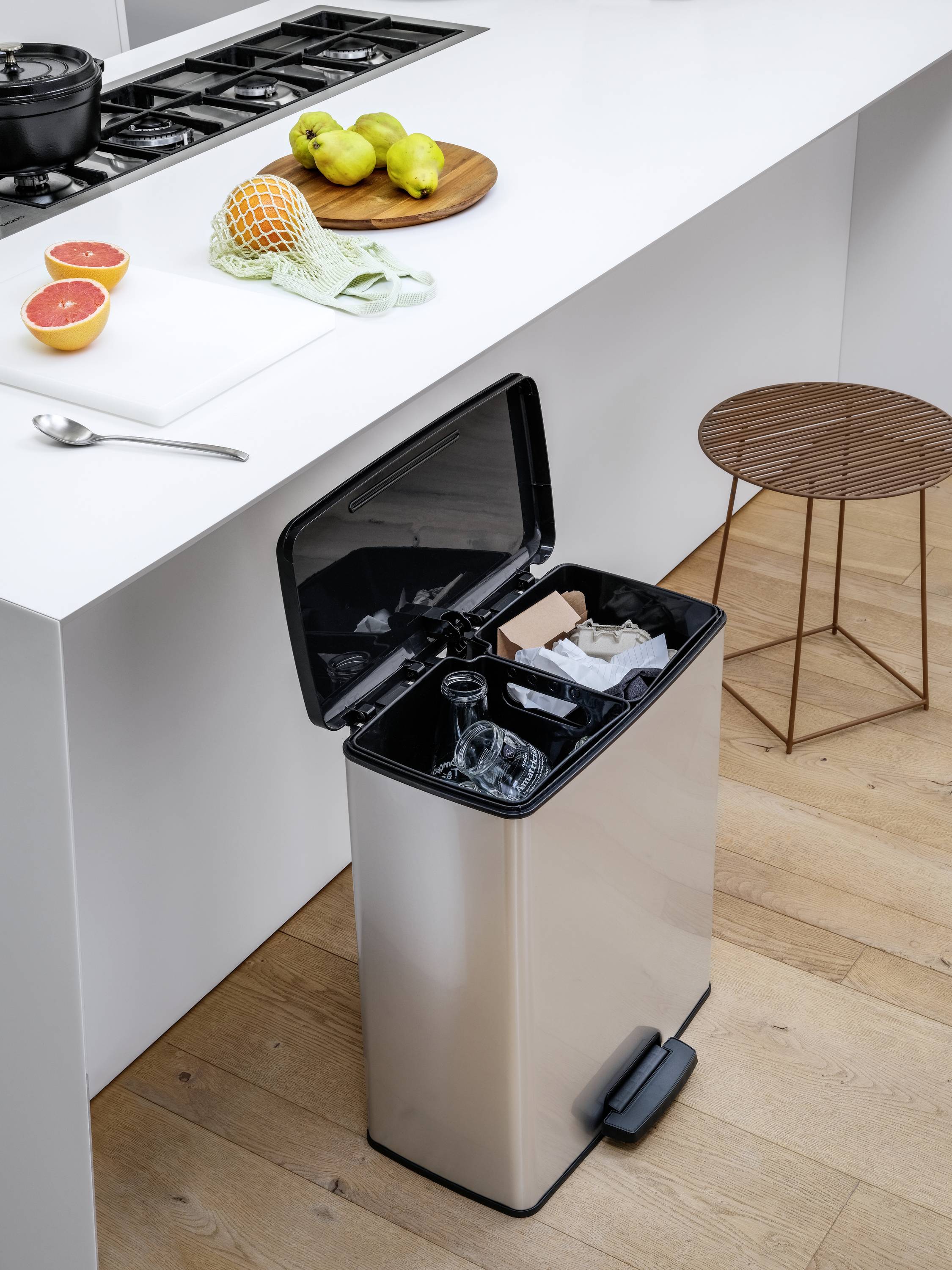 Une cuisine moderne avec un sol en bois clair. Sur l'îlot de cuisine, des fruits et une planche à découper sont posés. Une poubelle ouverte montre des bouteilles en verre vides.