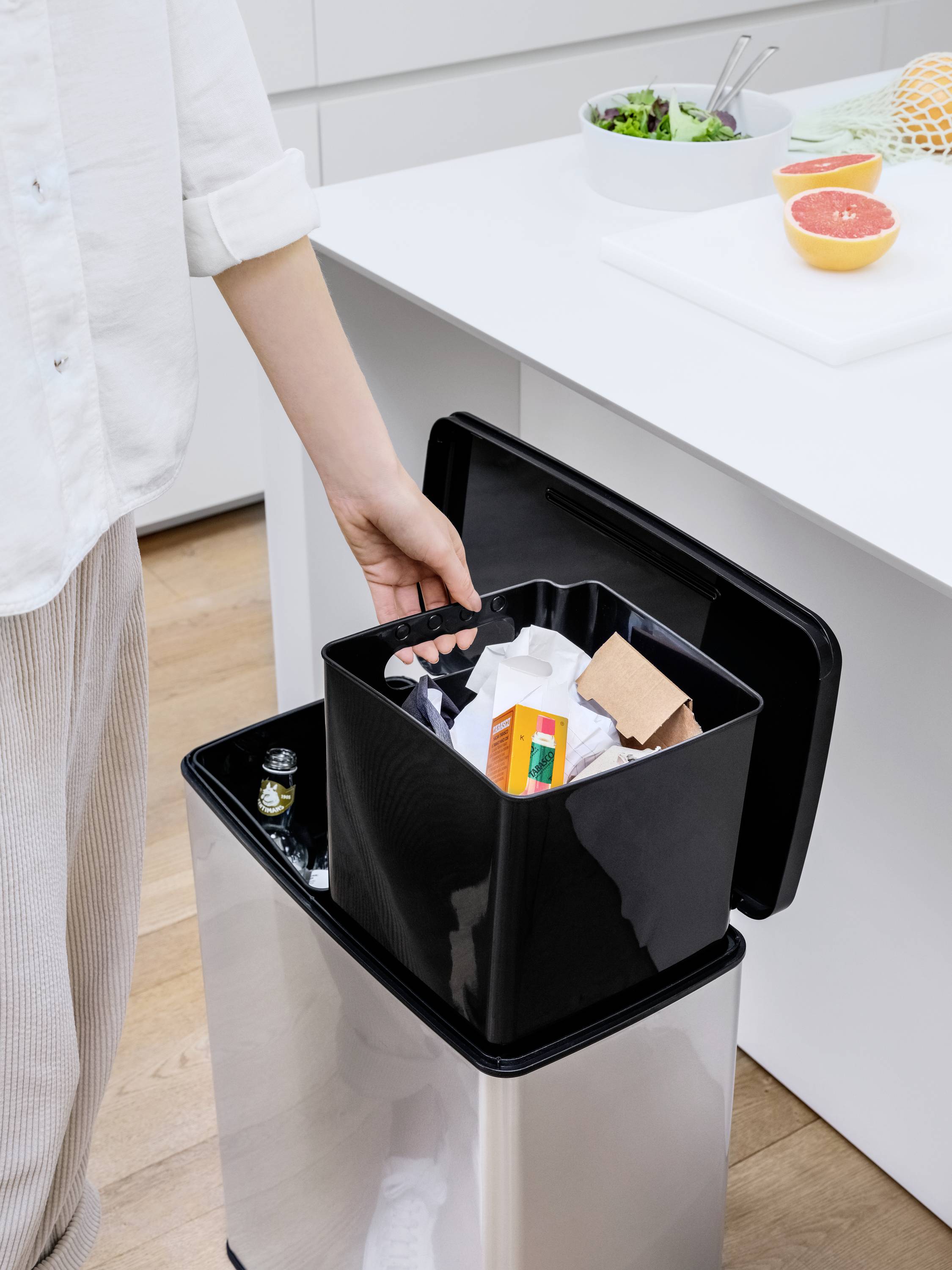 Une personne jette des déchets dans une poubelle dans une cuisine. Des fruits frais et une salade sont posés sur le plan de travail.