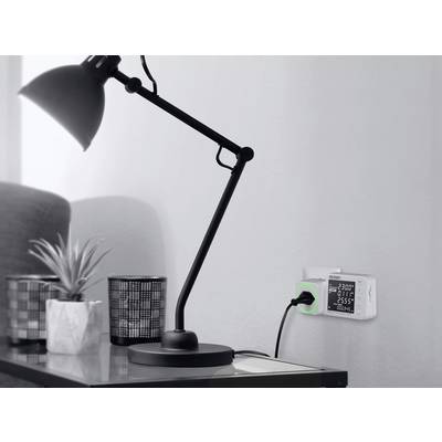 Une lampe de bureau à côté d'un vase et d'un gobelet. Un appareil de surveillance est encastré dans le mur, affichant des valeurs telles que la tension.