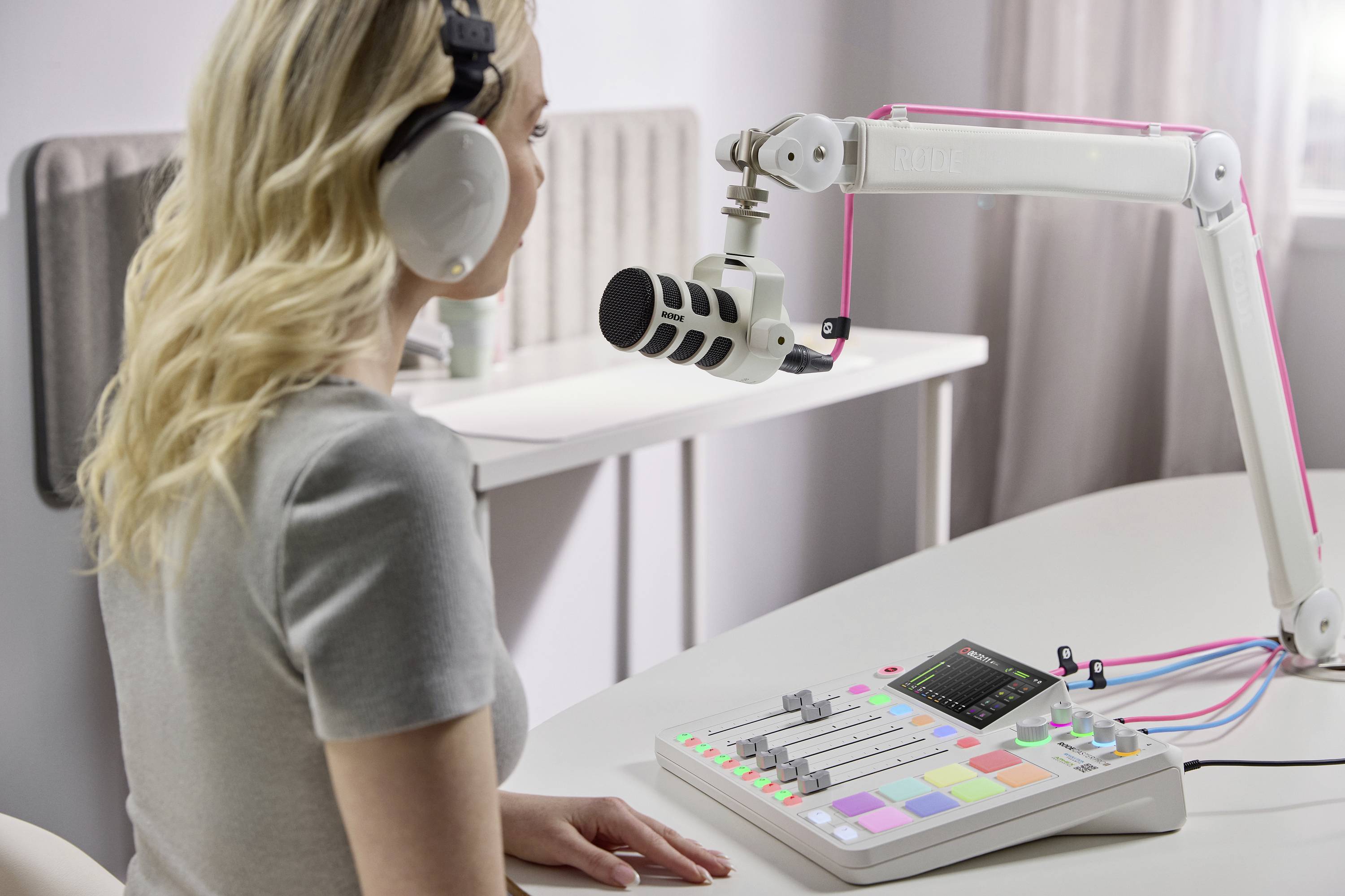 Une personne portant des écouteurs est assise devant un microphone et une console de mixage avec des touches colorées et un écran dans une pièce lumineuse.