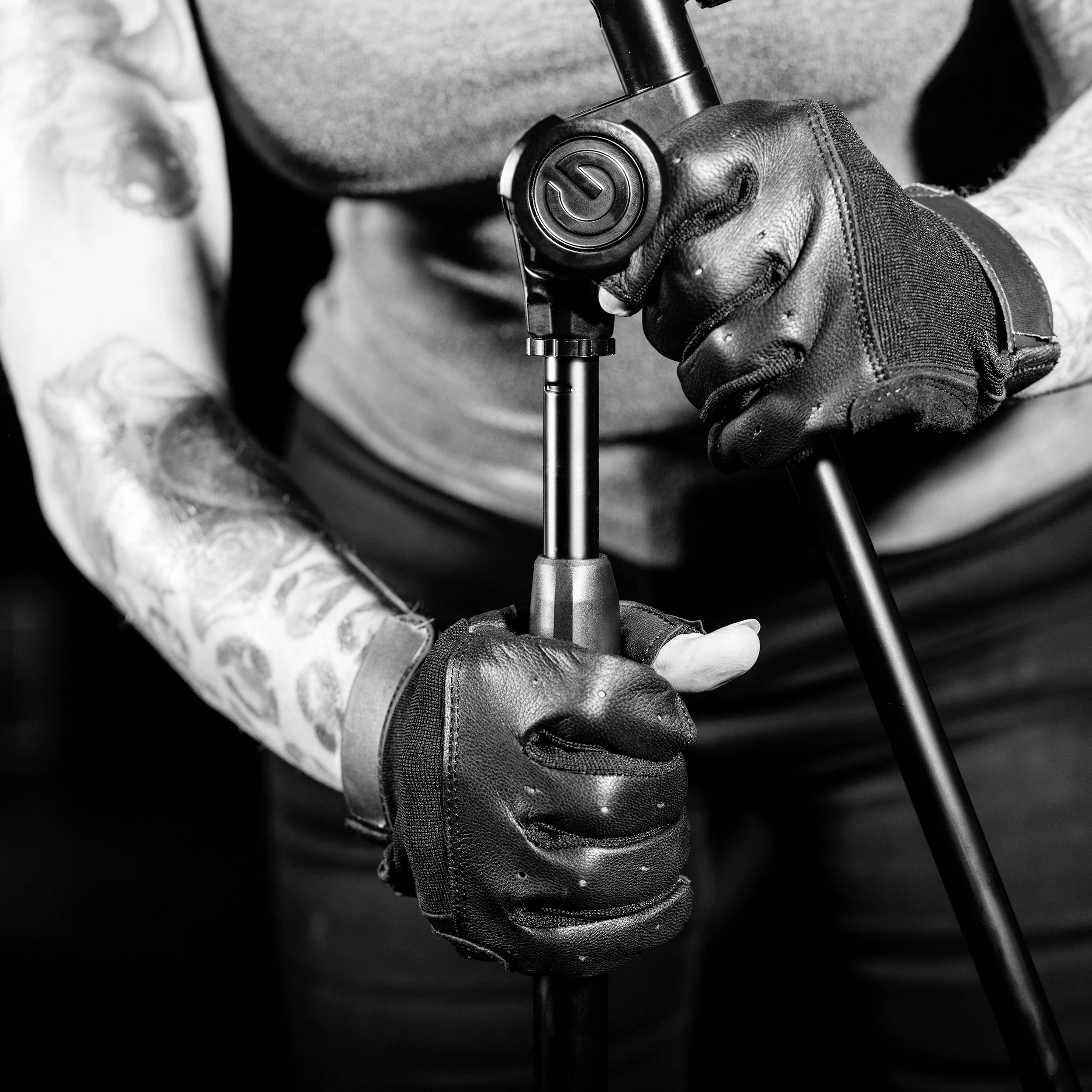 Une personne aux bras tatoués et portant des gants noirs tient un trépied dans une photographie en noir et blanc.