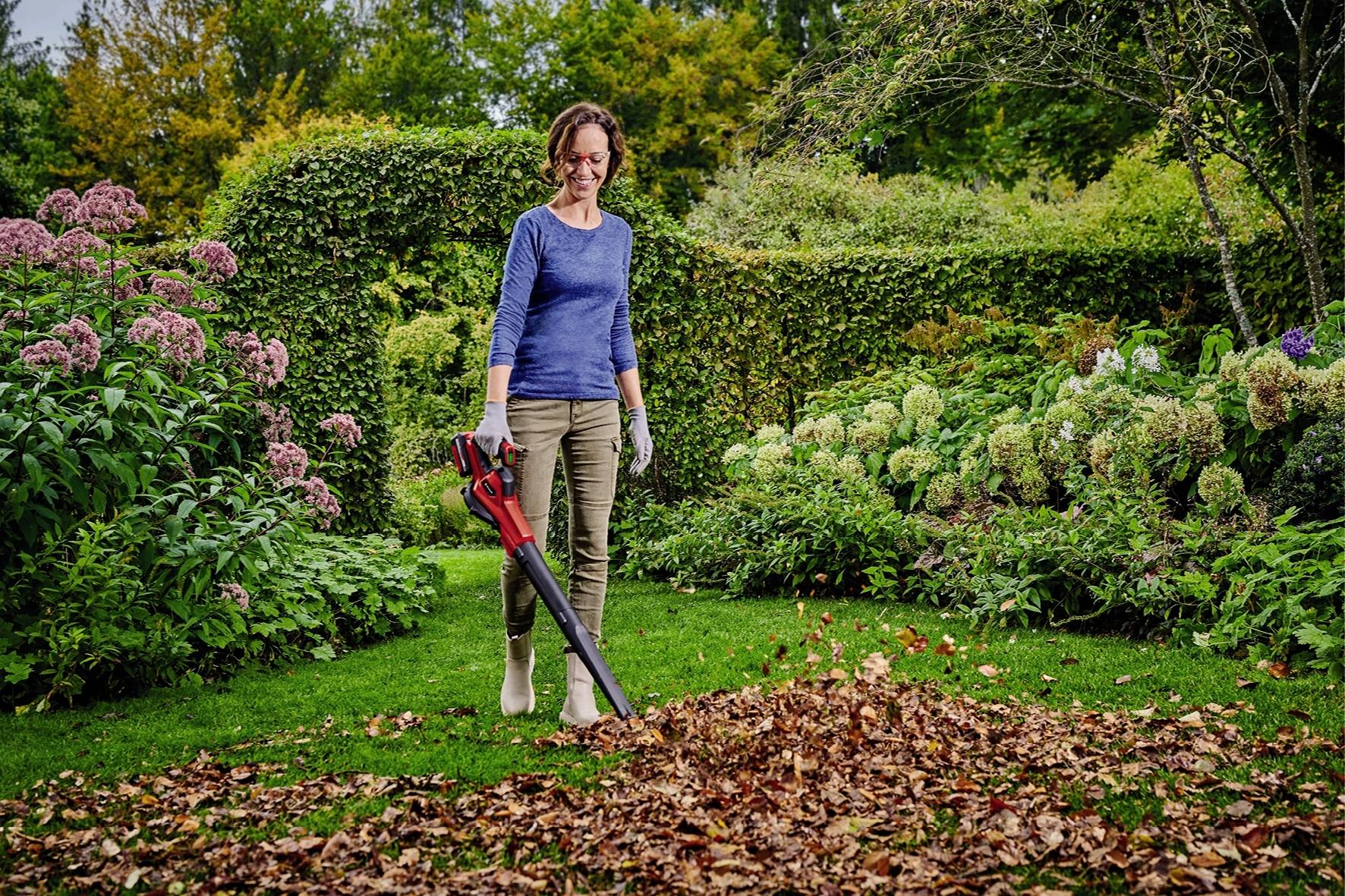 Une femme utilise un souffleur de feuilles pour enlever les feuilles sur une pelouse verte dans un jardin luxuriant avec des arbustes et des arbres.