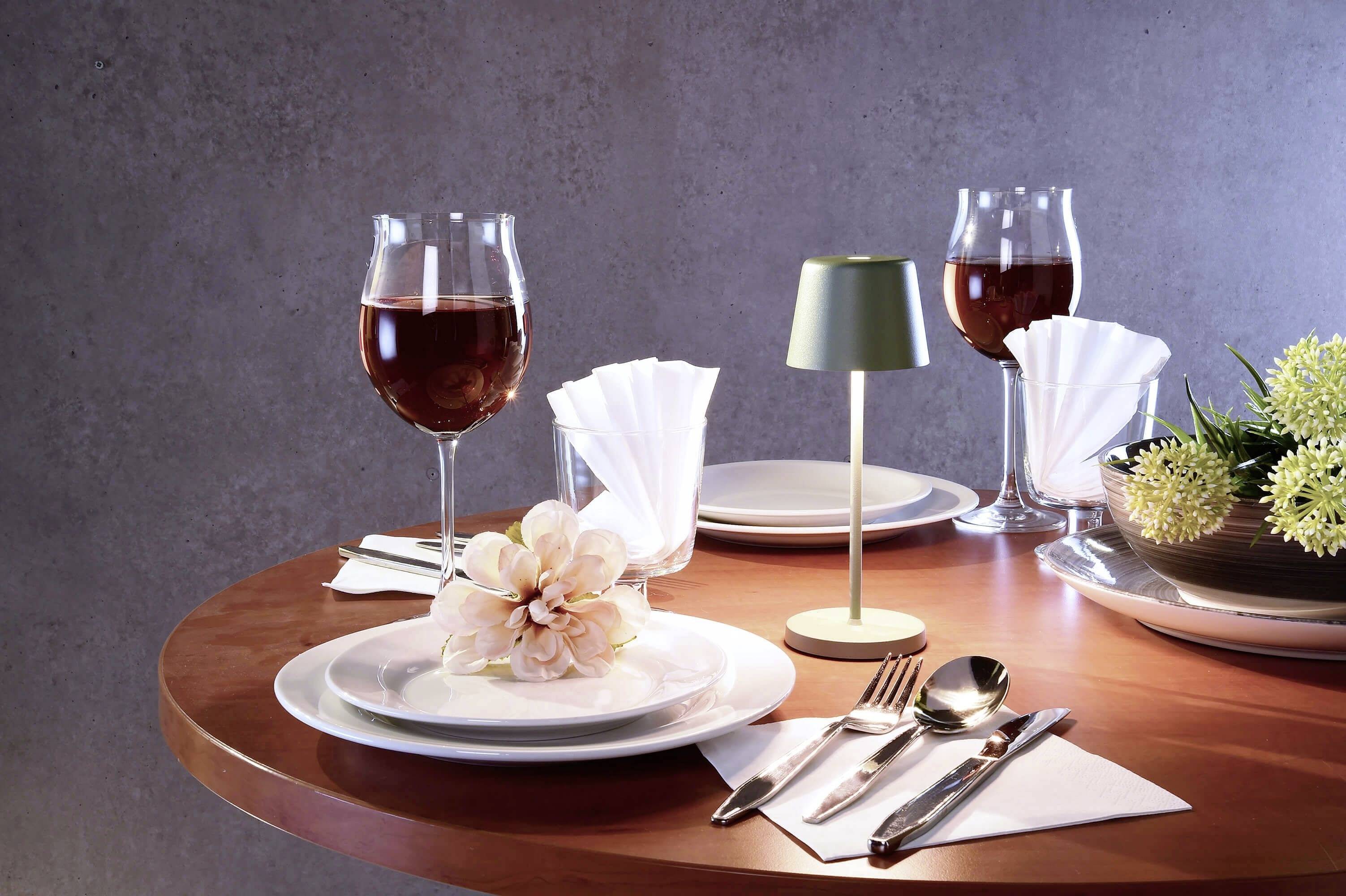 Une table ronde avec deux verres à vin, une fleur rose pâle sur une assiette, des couverts et une petite lampe de table.