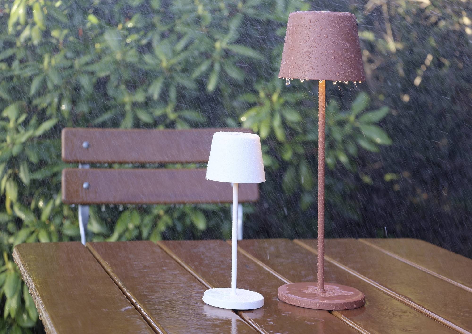 Deux lampes de table sous la pluie sur une table en bois, l'une blanche, l'autre brune. En arrière-plan : des plantes vertes et un banc en bois.