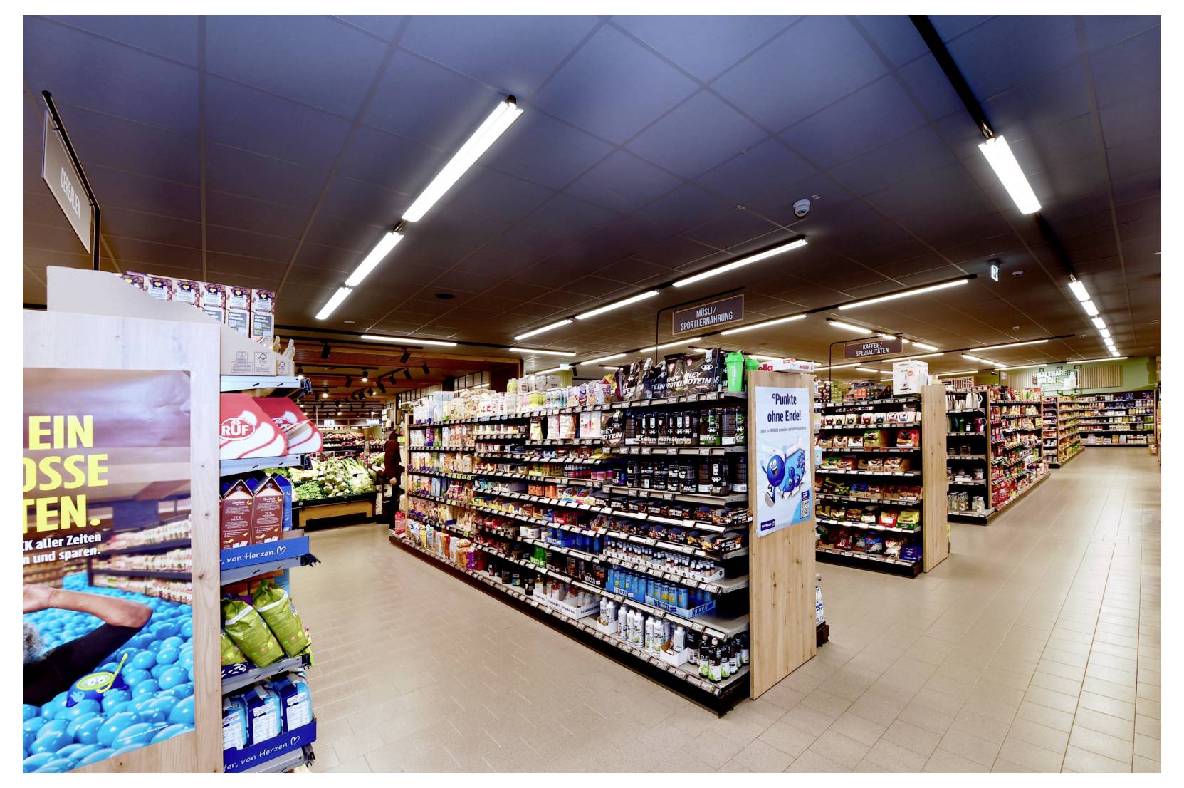 Vue en grand angle d'une allée de supermarché bien éclairée, avec des rayons garnis et des présentoirs de produits à l'intérieur d'une épicerie.
