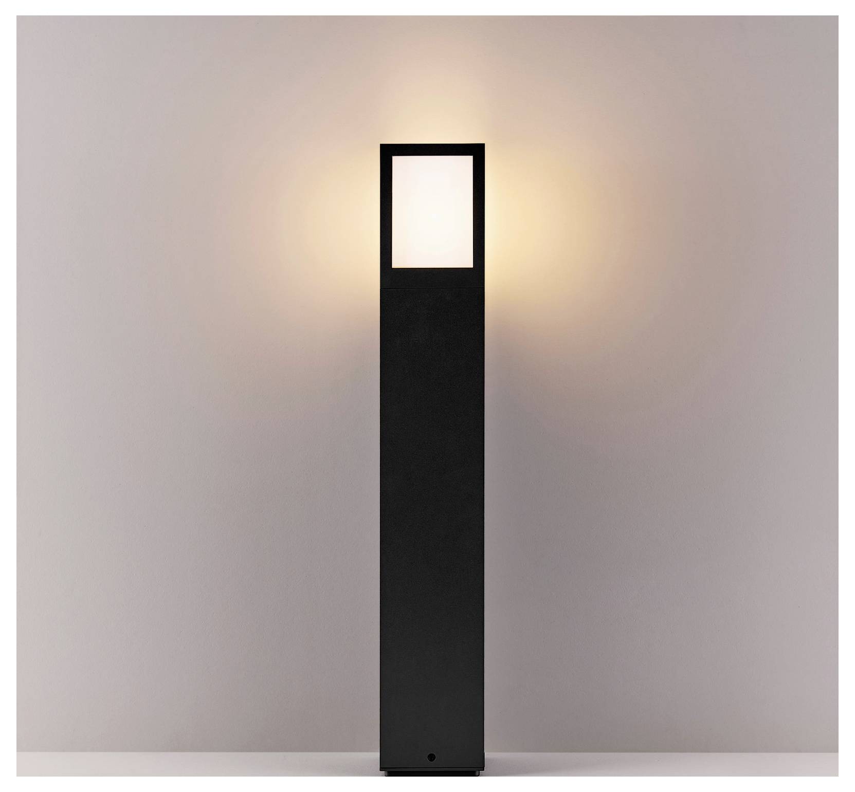 Grand lampadaire noir avec une lumière rectangulaire lumineuse en haut, devant un mur uni.