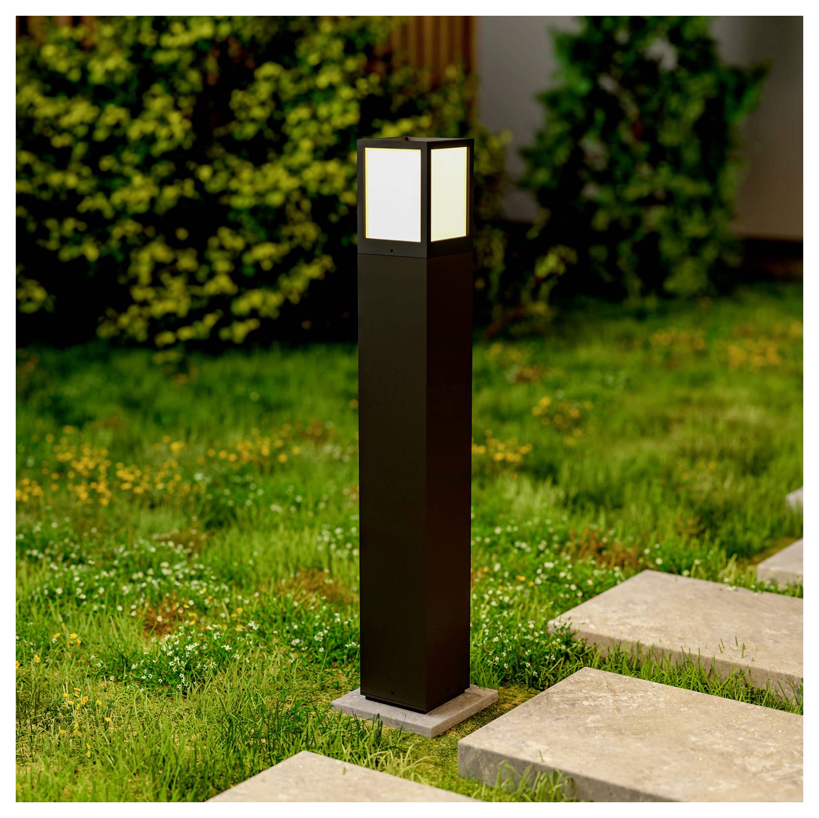 Applique de chemin d'extérieur moderne de couleur noire sur un socle en pierre, à côté d'une allée de jardin herbeuse, avec des panneaux carrés dépolis diffusant une lumière douce.