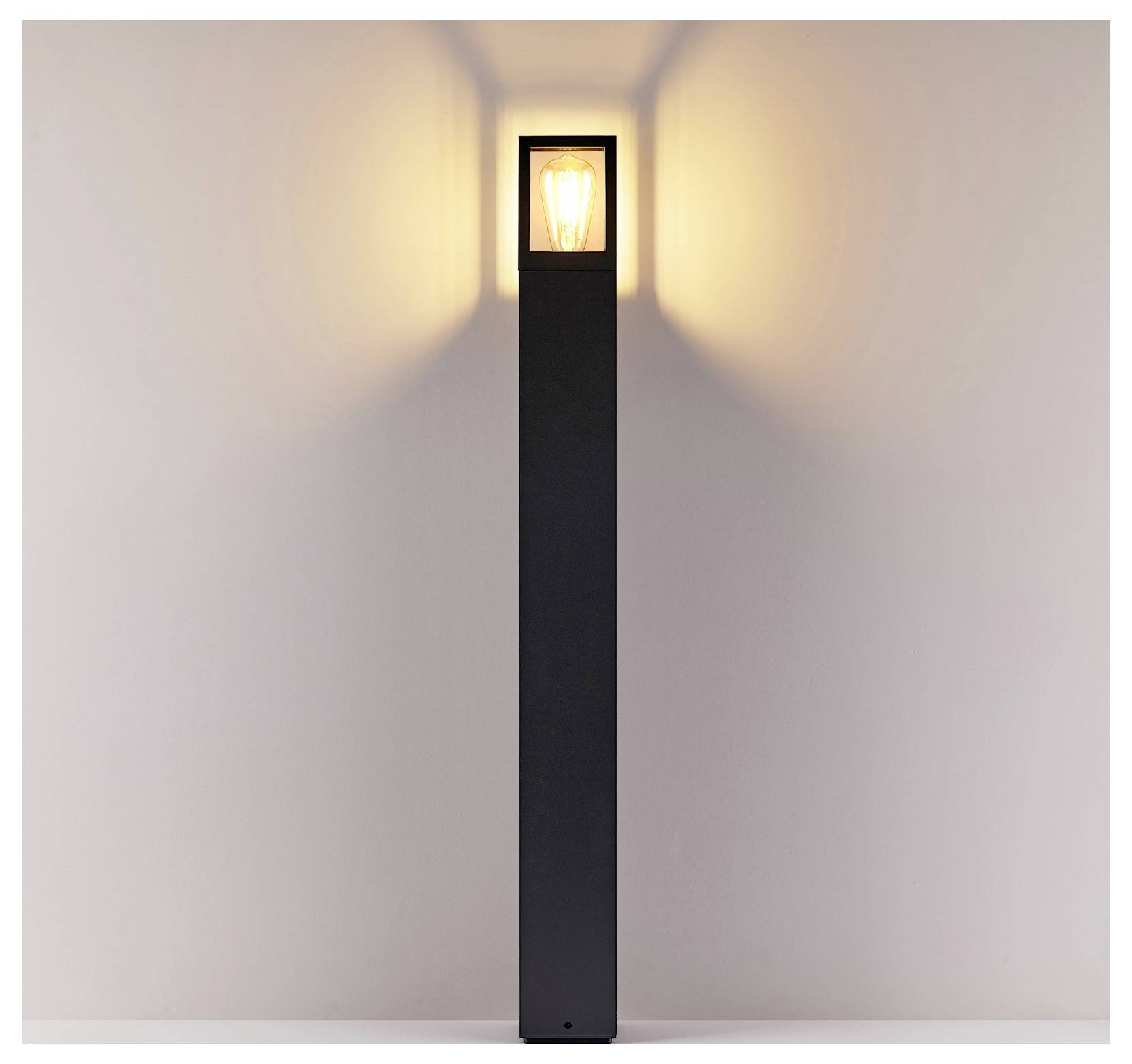 Applique d'extérieur noire et haute, fixée au mur, avec une ampoule lumineuse en haut, diffusant une lumière chaude sur le mur.