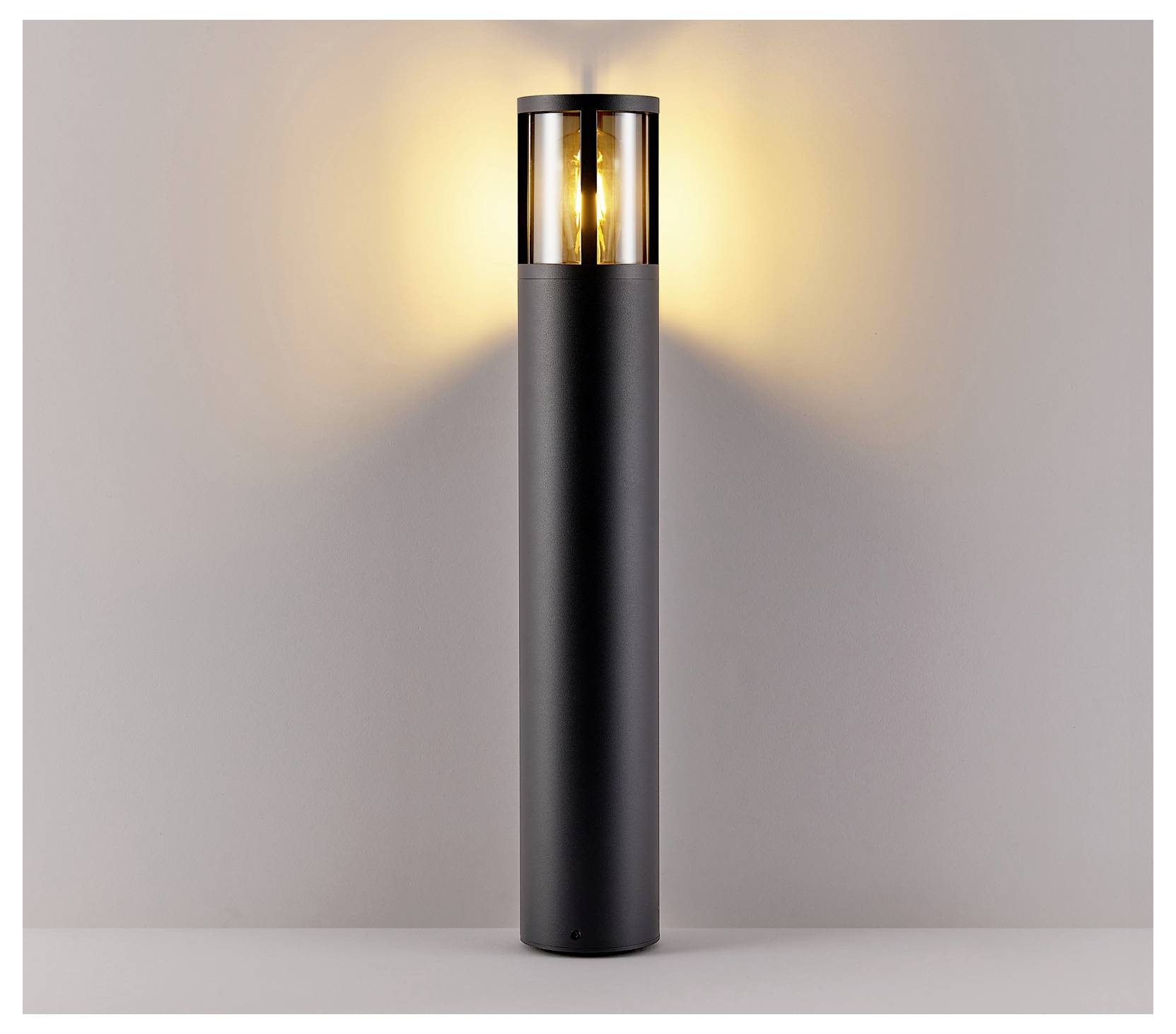 Borne d'éclairage extérieure cylindrique noire diffusant une lumière chaude contre un mur uni