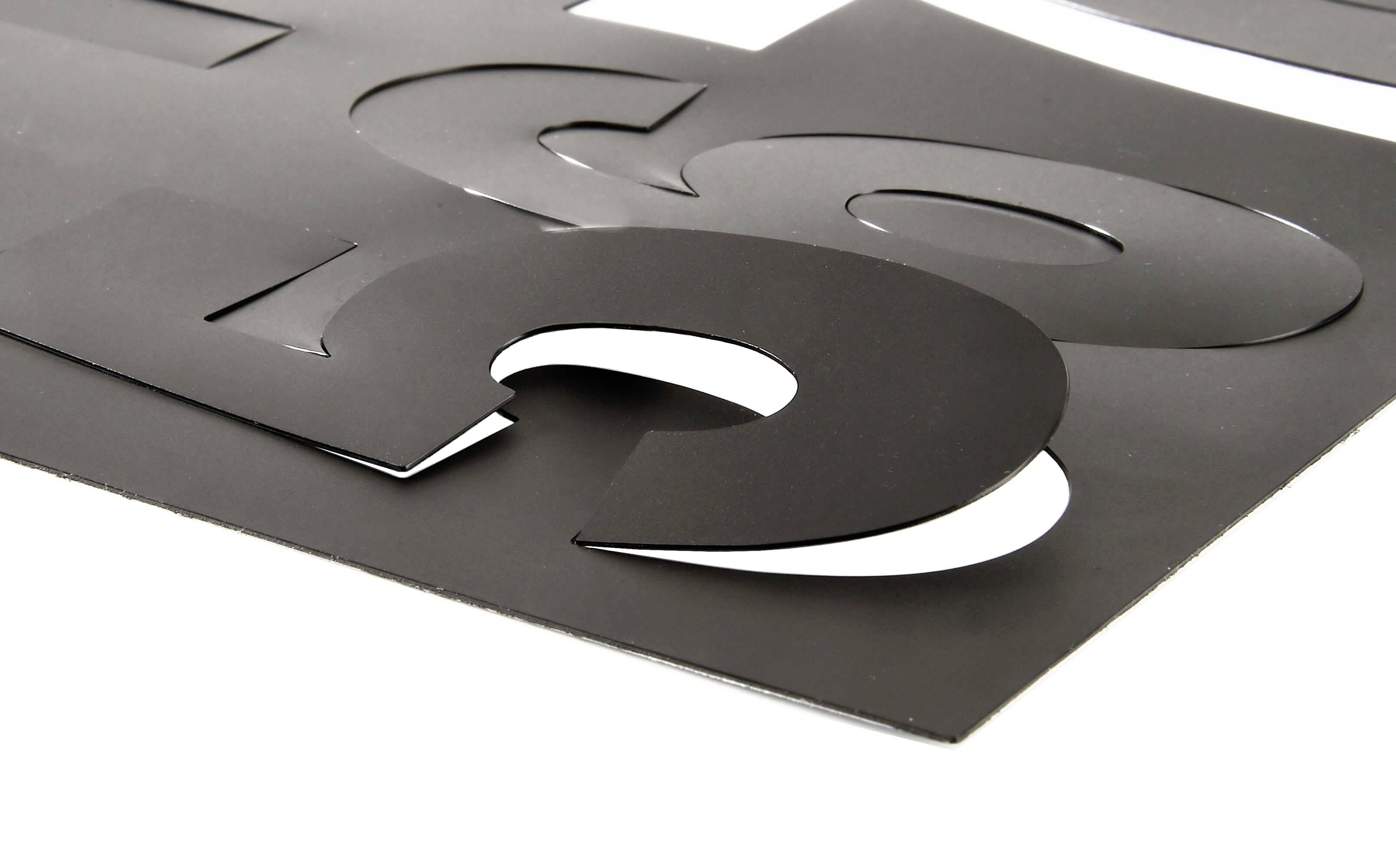 Des lettres noires découpées sur un fond blanc sont disposées de manière superposée sur une surface, créant un effet 3D.