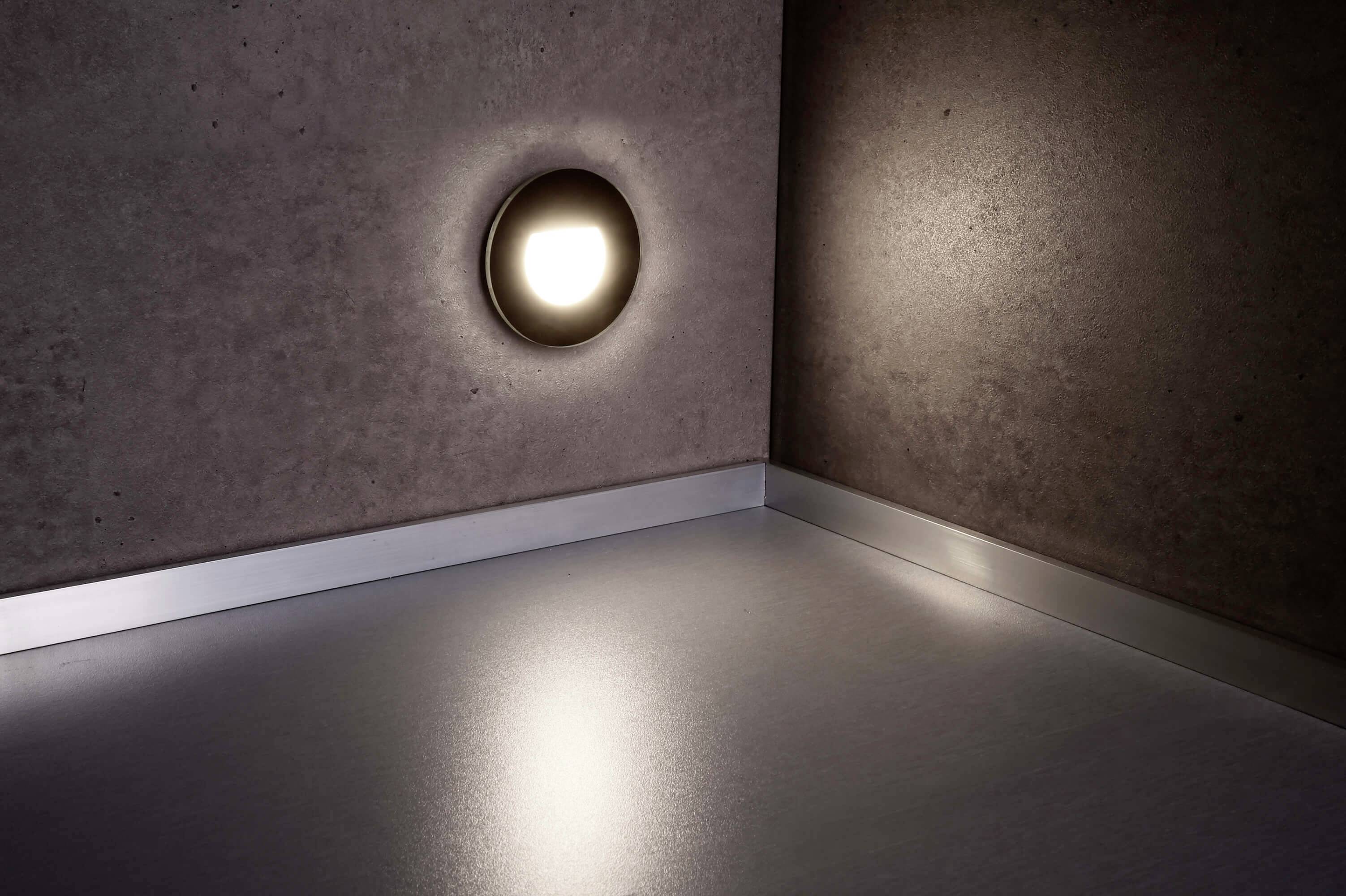 Un applique murale projette une lumière ronde et douce sur un mur en béton gris. Le sol reflète partiellement cette lumière.