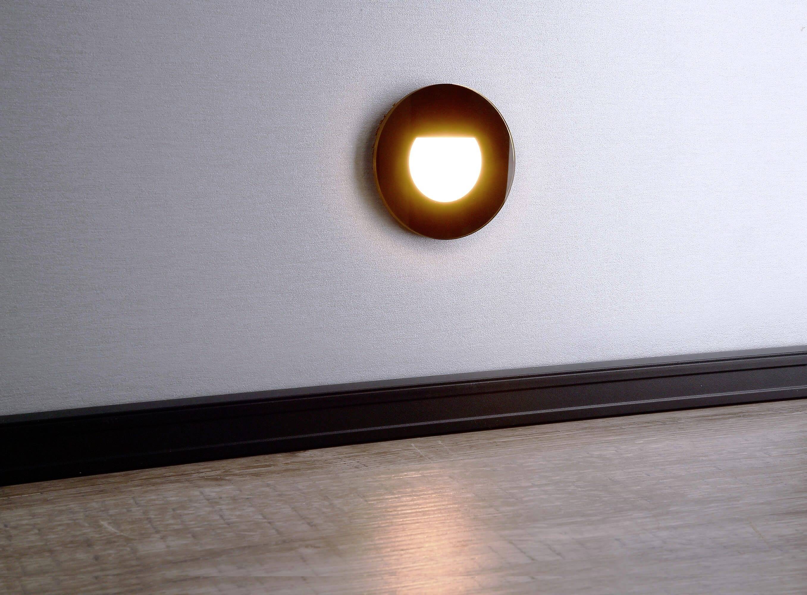 Un petit veilleuse LED ronde, encastrée dans le mur, projette une lumière douce sur un parquet en bois.