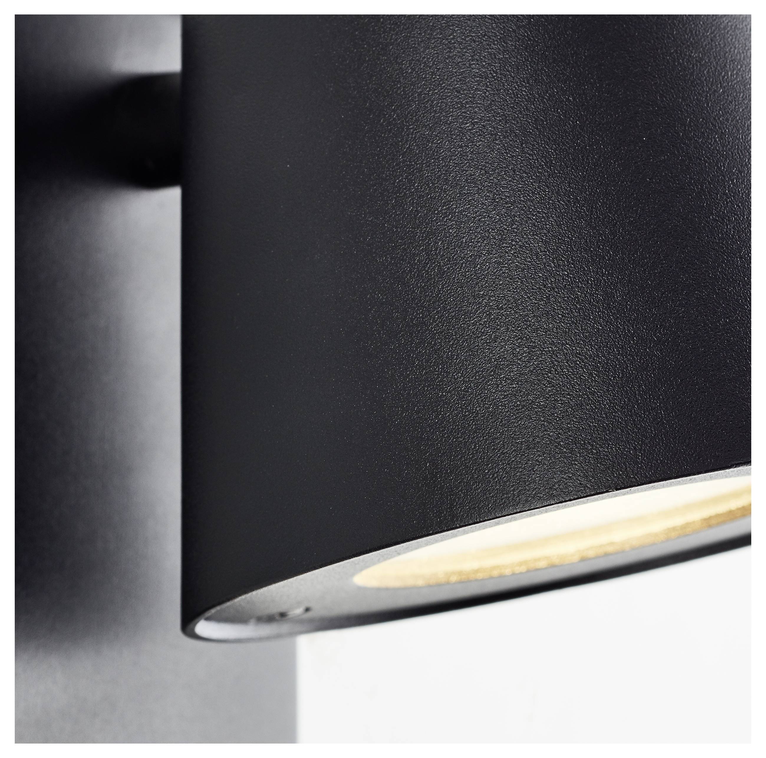 Un applique murale noir avec un abat-jour cylindrique diffuse une lumière chaleureuse. Elle est montée sur un mur clair.