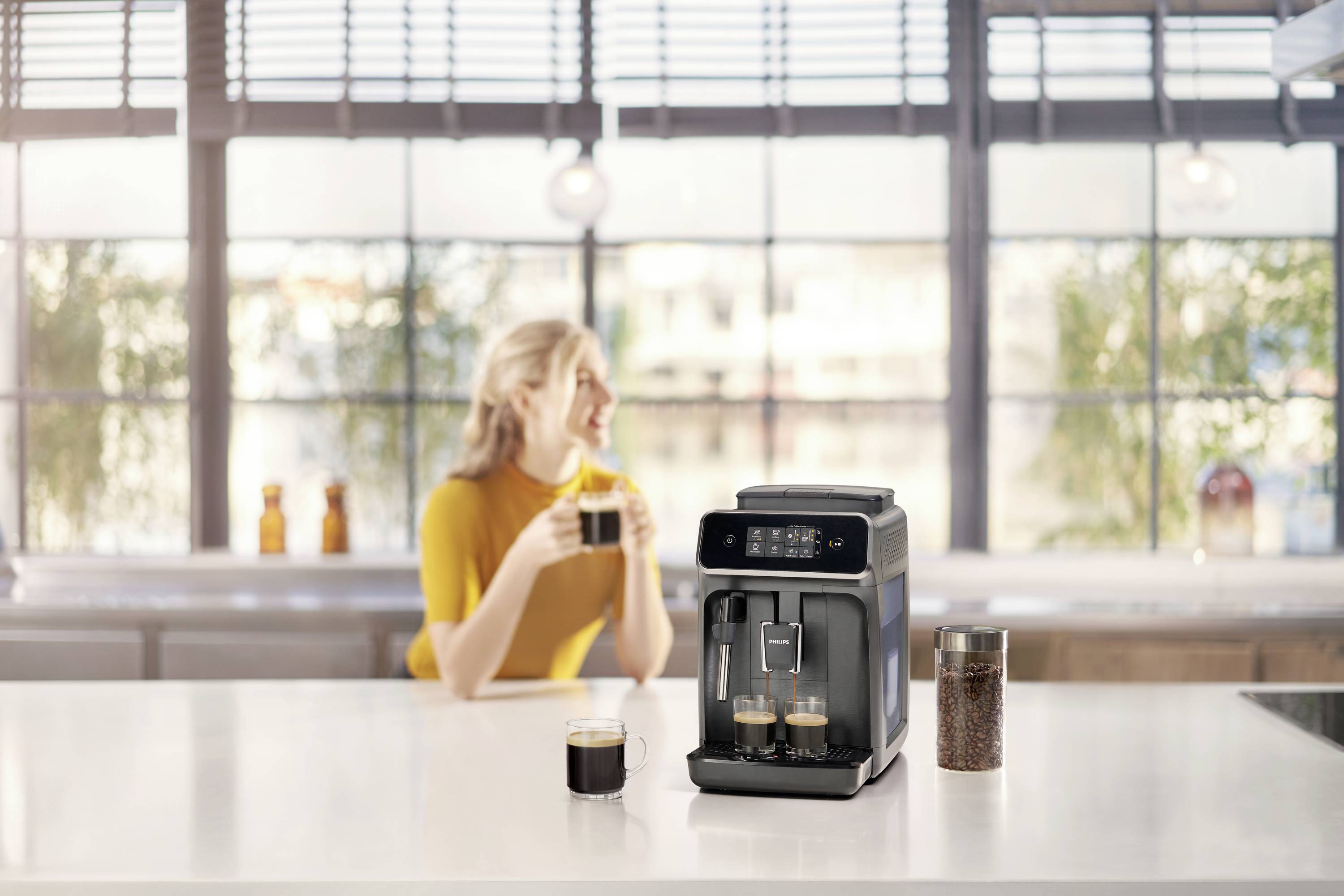 Une femme est assise dans une cuisine et tient une tasse de café. À côté d'elle, une machine à café moderne est posée sur le plan de travail.