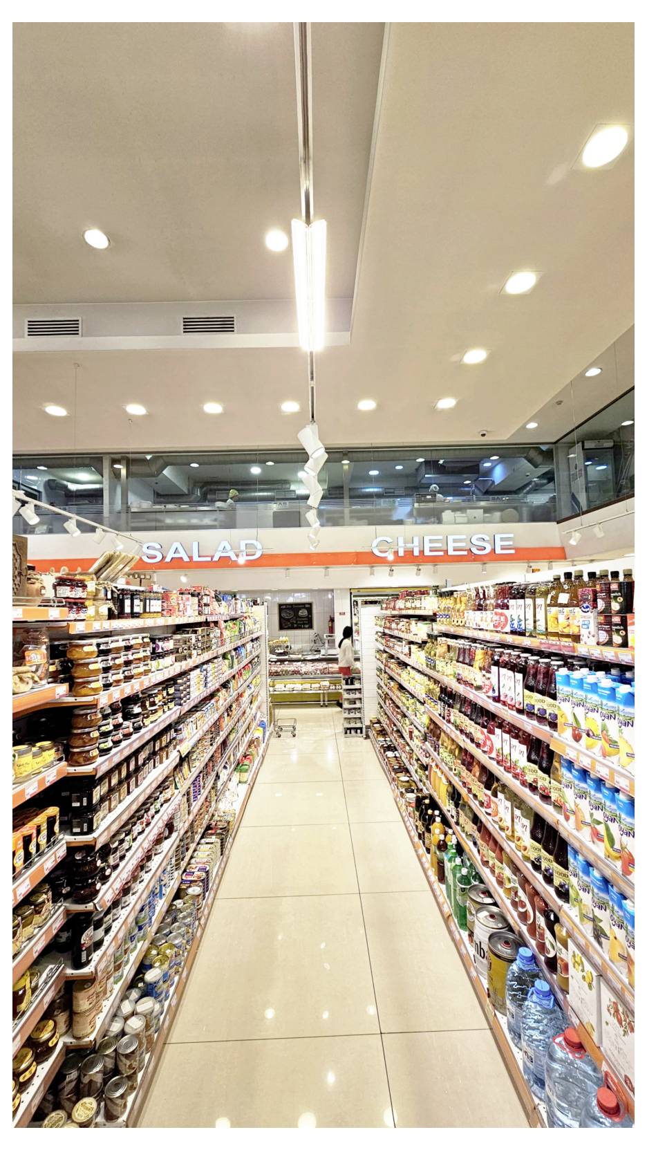 Allée de supermarché avec des rayons de produits emballés et de boissons, menant à un présentoir du fond sous les panneaux « SALAD » et « CHEESE ».