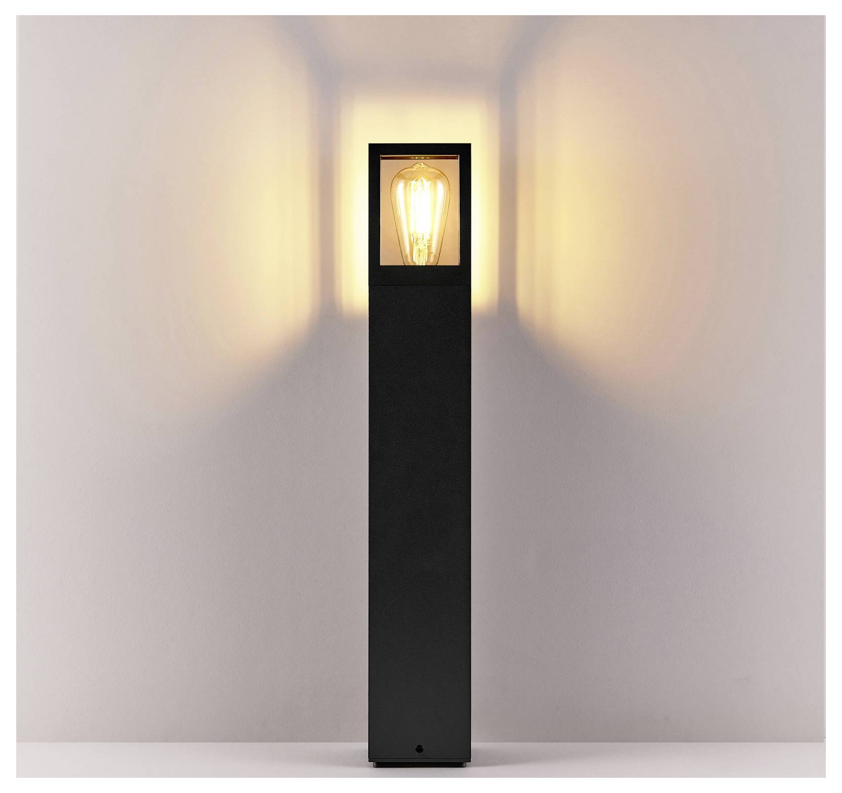 Grand lampadaire noir avec une ampoule nue lumineuse, diffusant une lumière chaude sur un mur uni.