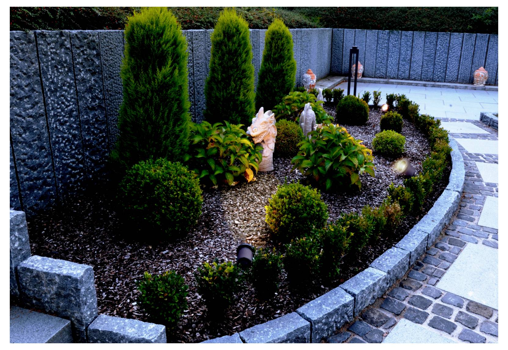 Petit parterre paysager avec arbustes, grands conifères à feuillage persistant, bordure en pierre et allée longeant une clôture grise.