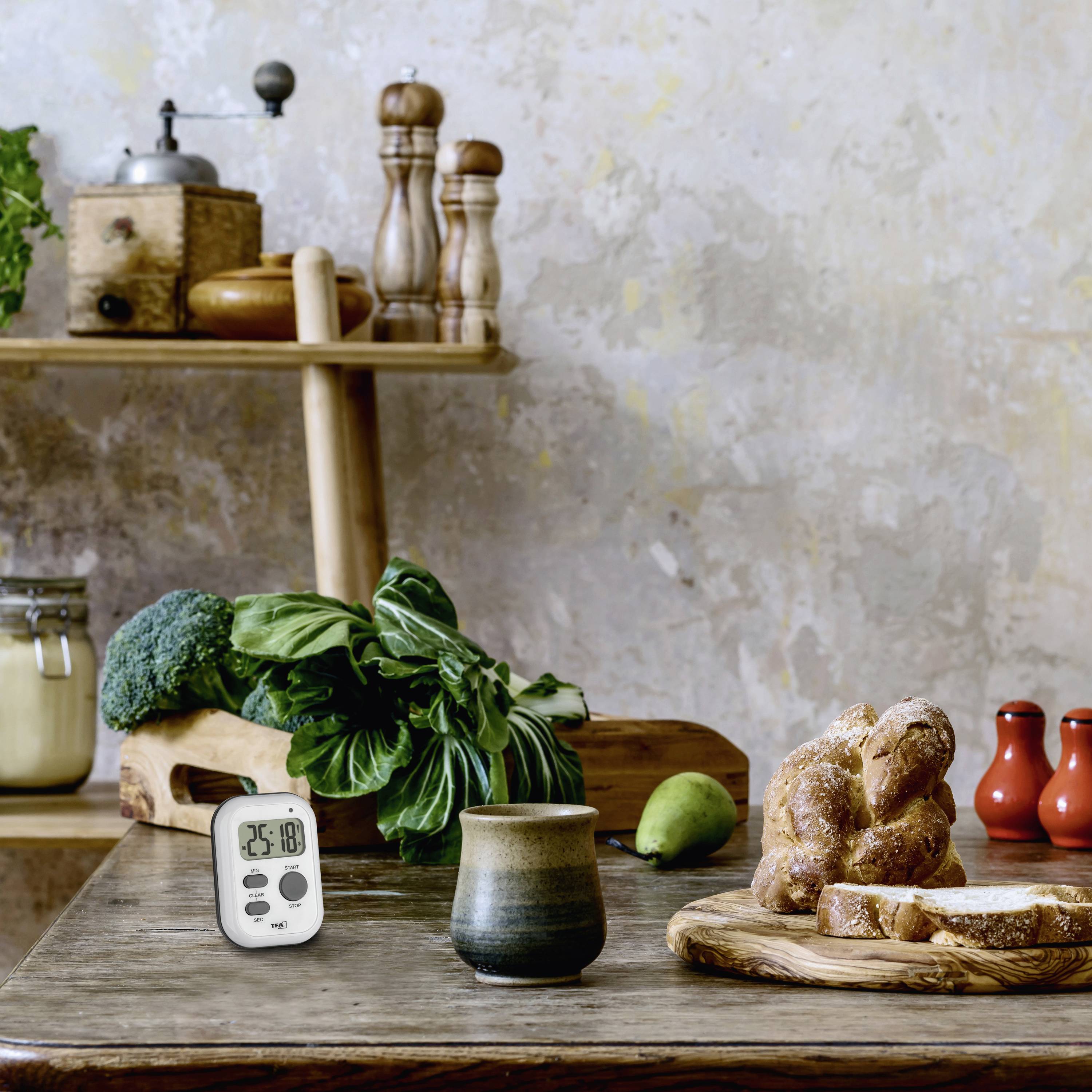 Une table avec du pain frais, du brocoli, un minuteur et une tasse, avec en arrière-plan un environnement de cuisine rustique et des moulins à épices.
