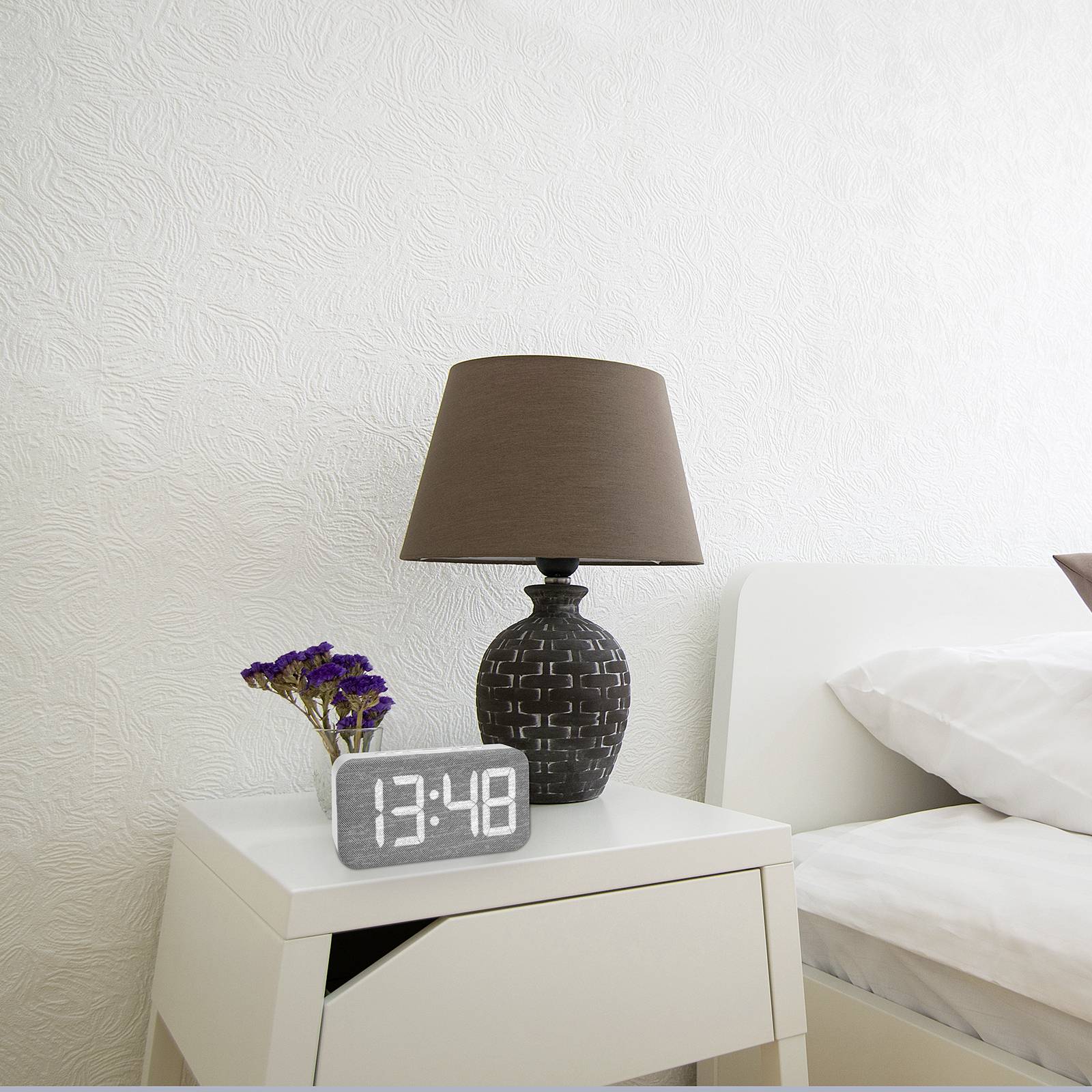 Lit avec des draps blancs, à côté une table de chevet avec une horloge numérique (13:48), une lampe de table et des fleurs séchées dans un vase.
