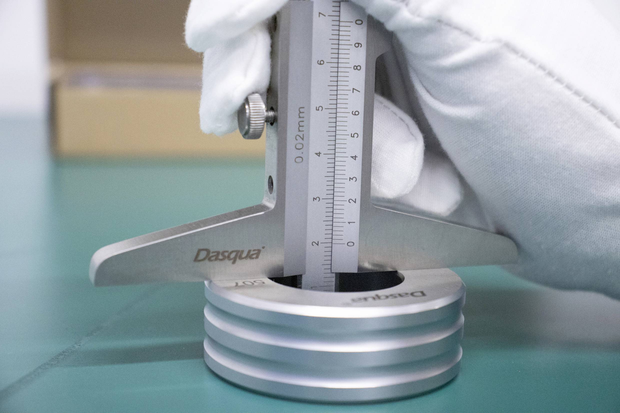 Une main gantée de blanc tient un pied à coulisse et mesure l'épaisseur d'un objet cylindrique métallique posé sur une table.