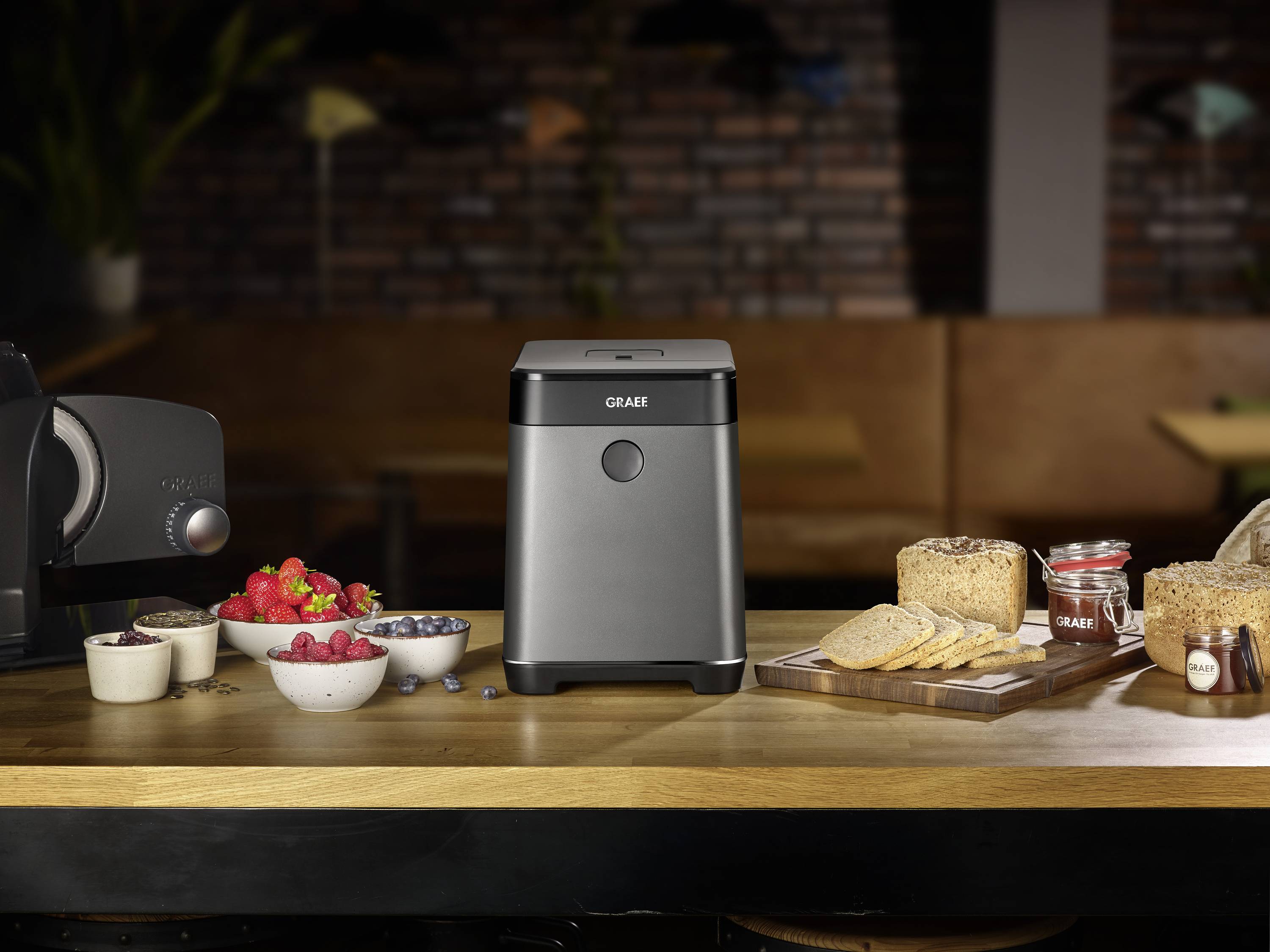 Une machine à pain est posée sur une table en bois dans une cuisine. Elle est entourée de différents types de pain, de fraises fraîches et de confiture.