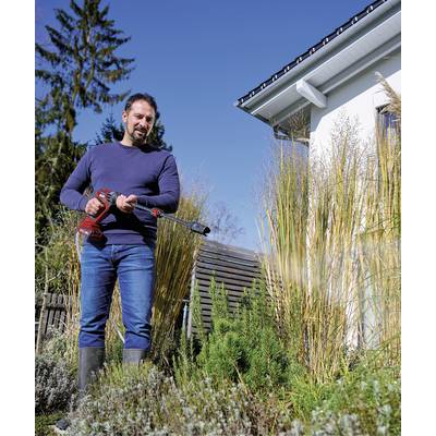 Un homme en jeans et pull coupe des plantes dans le jardin avec un taille-bordures électrique. Une maison est visible en arrière-plan.