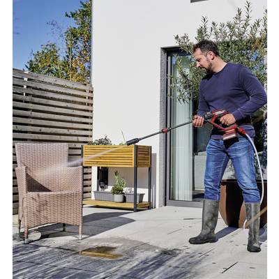 Un homme nettoie une chaise tressée avec un nettoyeur haute pression sur une terrasse. Des plantes et des meubles sont visibles en arrière-plan.