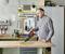 Un homme utilise un aspirateur à main pour enlever des copeaux de bois d'une table dans un atelier. Des outils sont suspendus au mur.