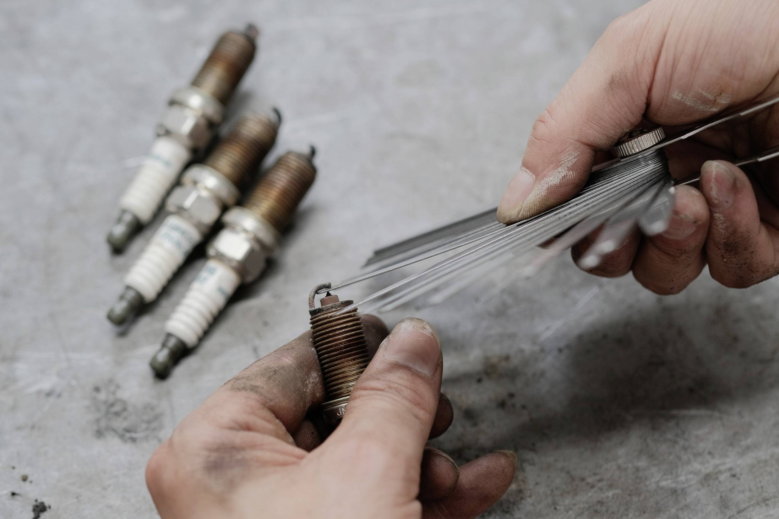 Une personne mesure l'écartement des électrodes d'une bougie d'allumage à l'aide d'une jauge d'épaisseur. En arrière-plan, plusieurs bougies d'allumage usagées sont posées.