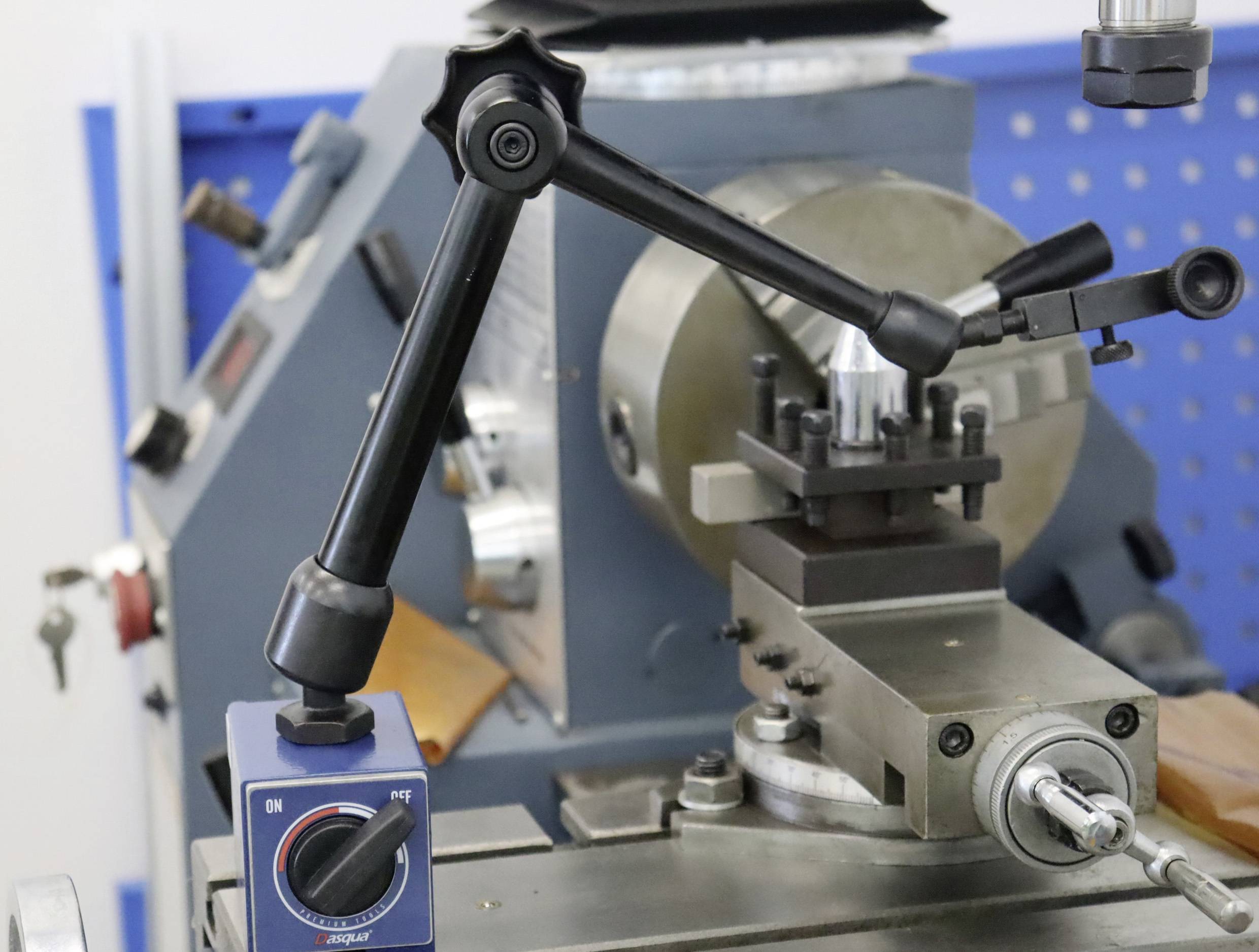 Une machine-outil de tournage en métal avec un mandrin de serrage, un porte-outil et un bras de rotation. L'arrière-plan montre un mur d'outils.