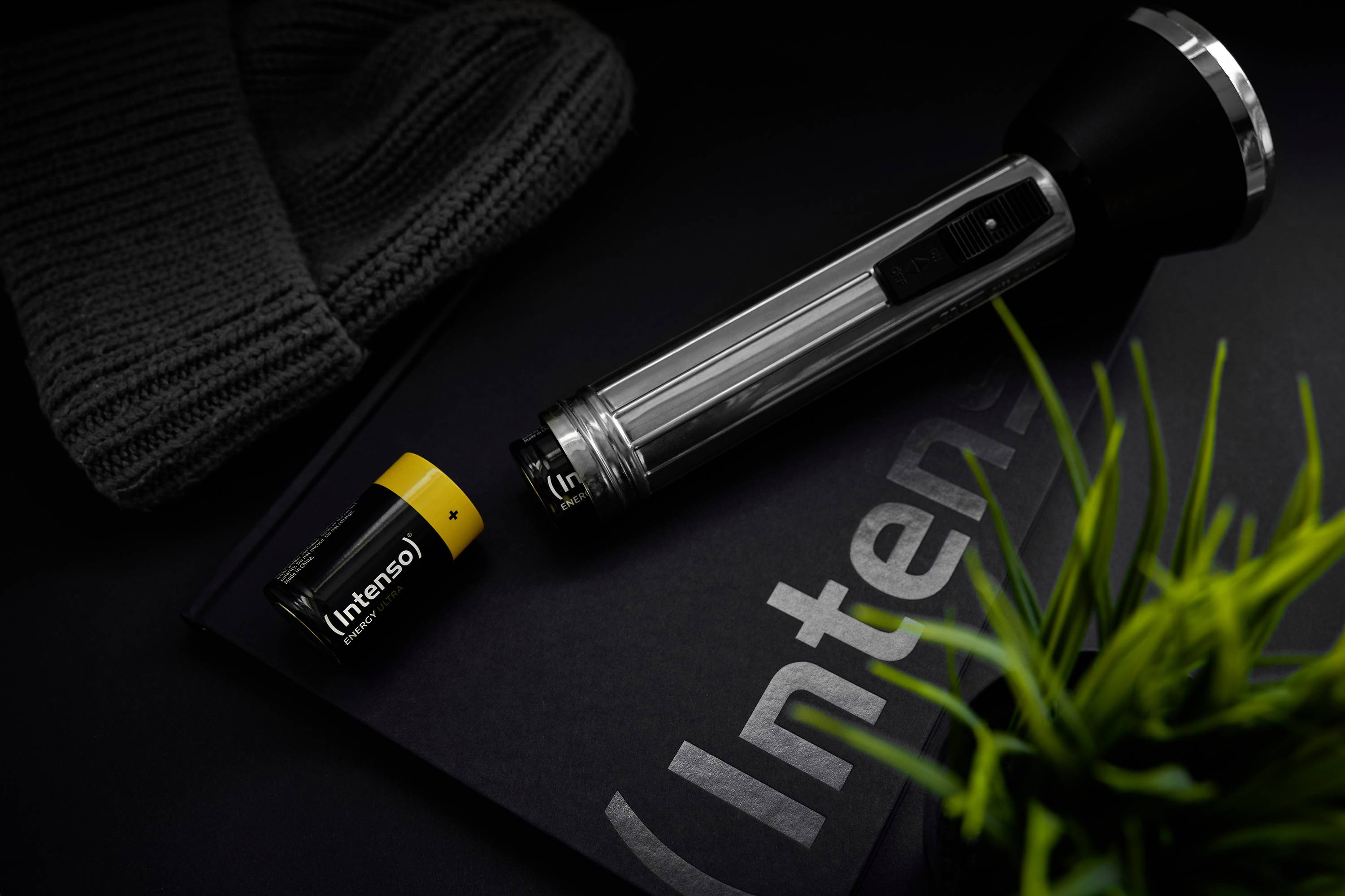 Une lampe de poche noire et une batterie jaune de la marque 'Intenso' sont posées sur un fond sombre à côté d'un chapeau gris.