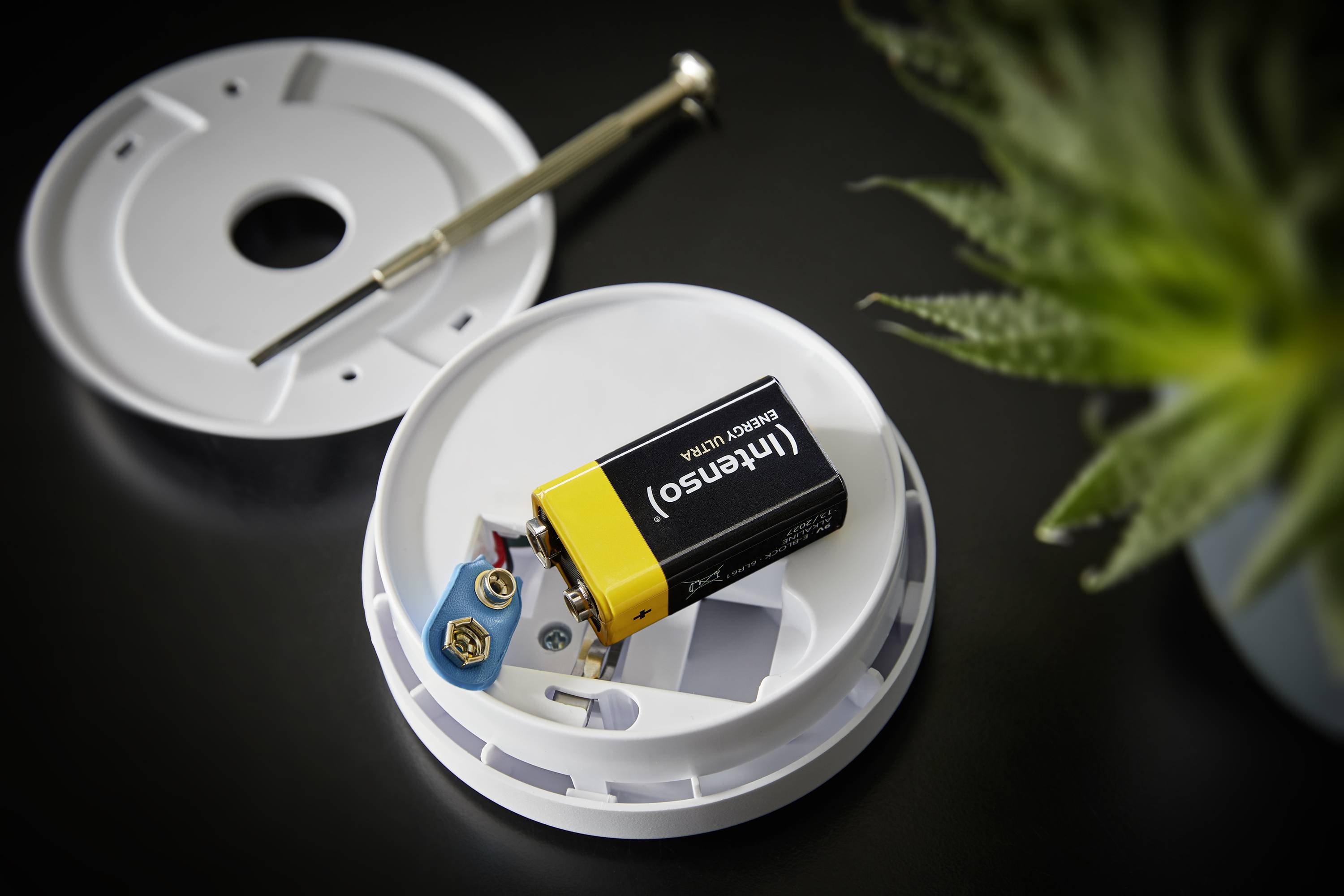 Un détecteur de fumée ouvert avec son couvercle retiré, montrant une batterie et des connexions câblées, avec un tournevis posé à côté sur la table.