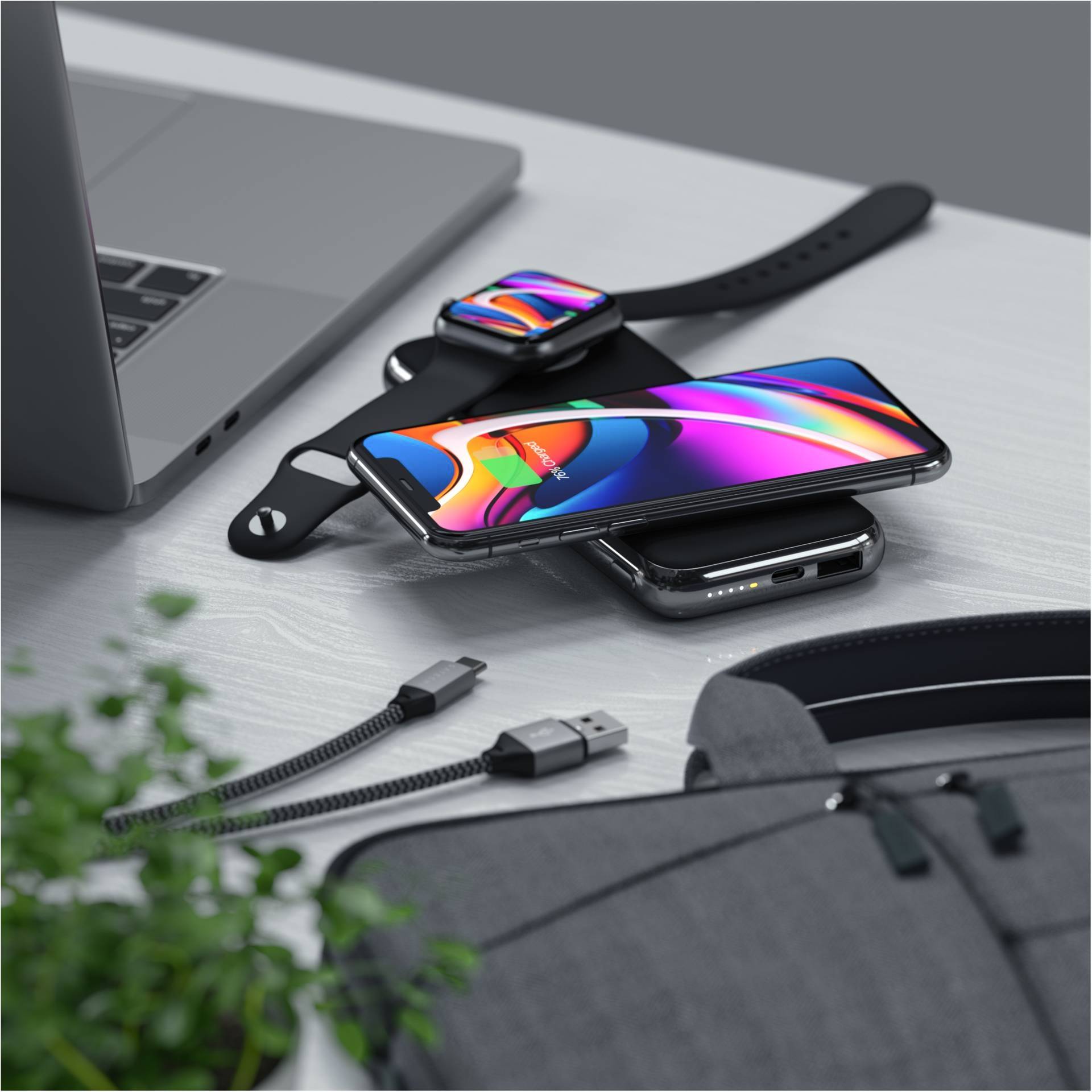 Un smartphone, une smartwatch et un ordinateur portable sont posés sur une table. Le smartphone est en train d'être chargé sans fil. Des câbles USB et un sac gris sont visibles.