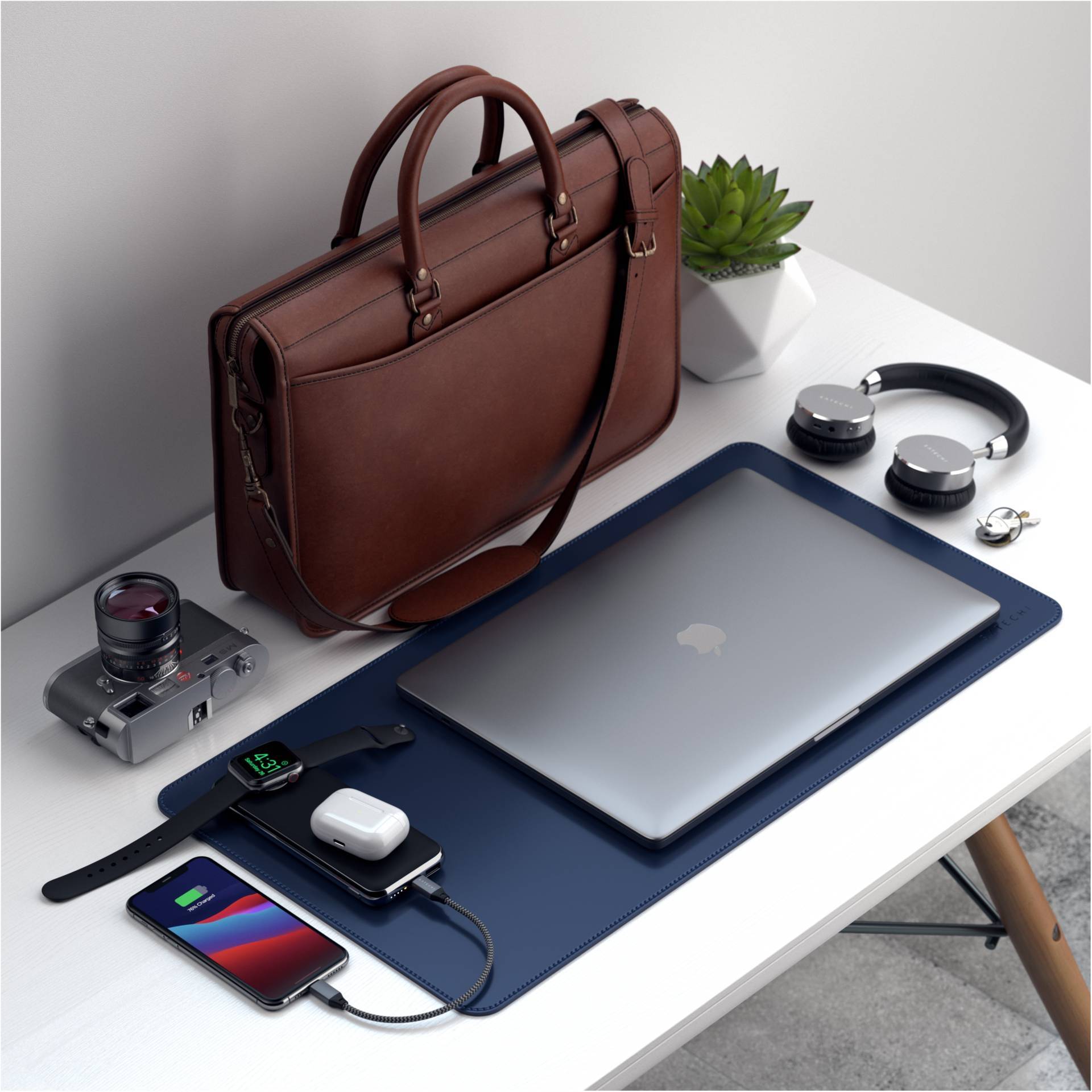 Sur un bureau sont posés un ordinateur portable, un sac en cuir, un appareil photo, des écouteurs, un smartphone, une montre connectée et des plantes.