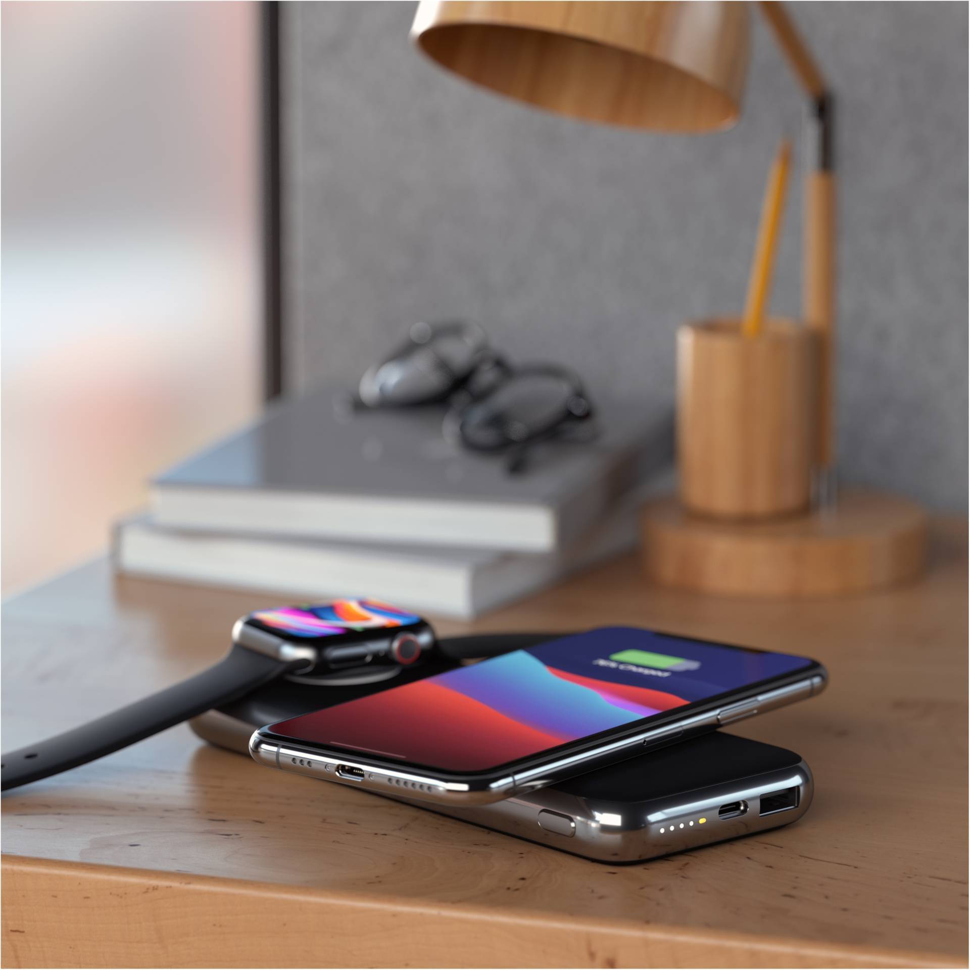 Un smartphone et une smartwatch se chargent sans fil sur une table, à côté desquels sont posés des lunettes et des livres. Une lampe se trouve en arrière-plan.
