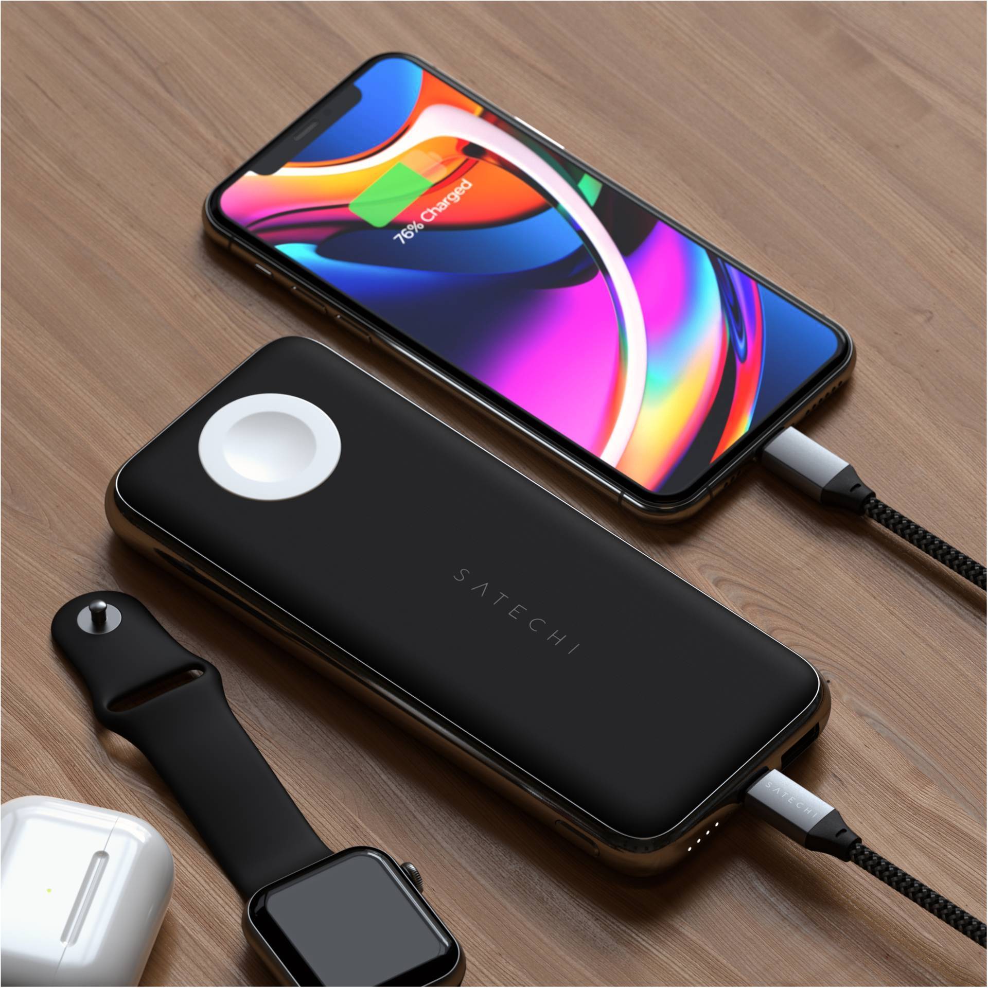 Smartphone et chargeur multifonction sur une table en bois. Le smartphone affiche "76% chargé". Une montre connectée est en charge à côté des AirPods.