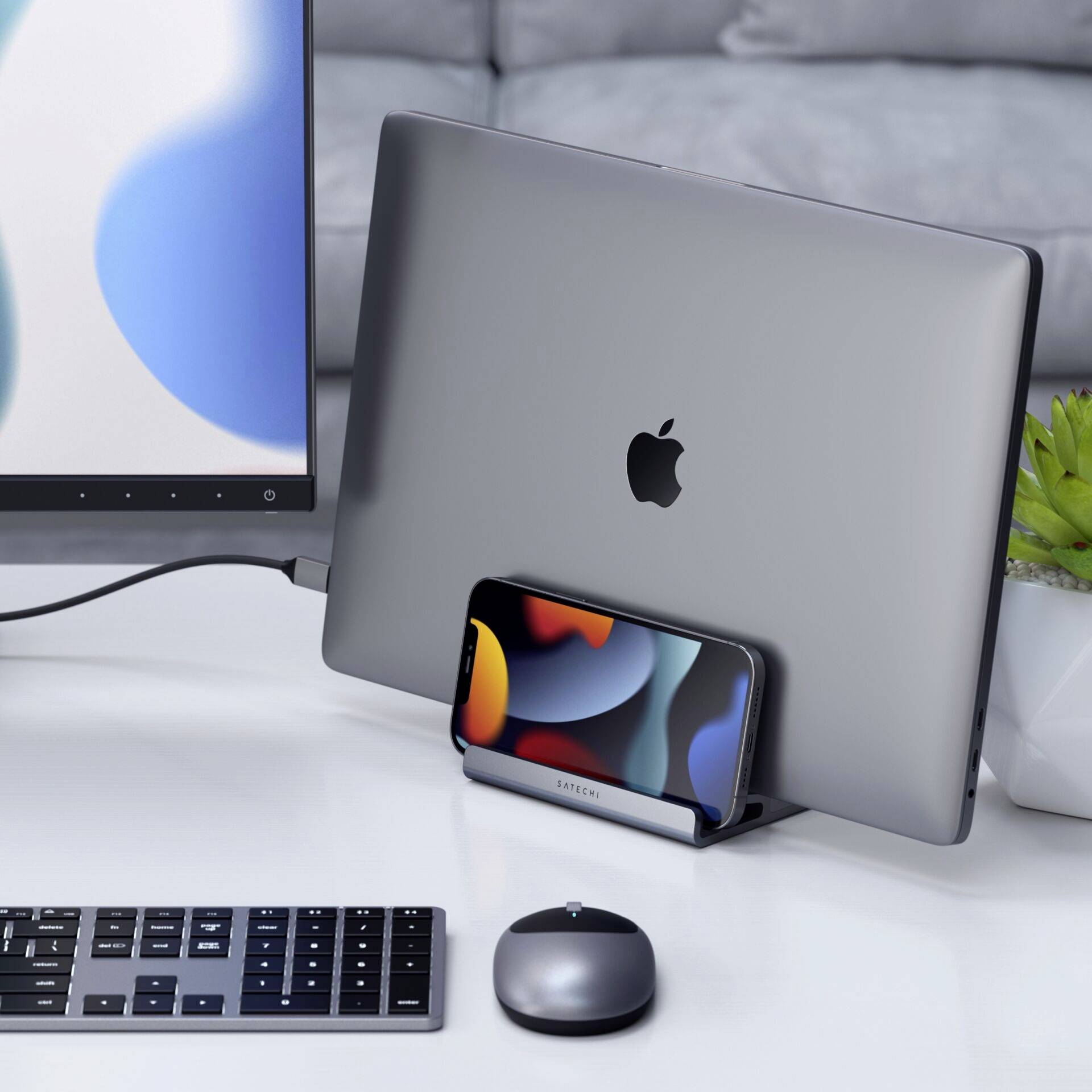 Un poste de travail à domicile avec un ordinateur portable, un smartphone dans un support, un clavier et une souris sur une table blanche.