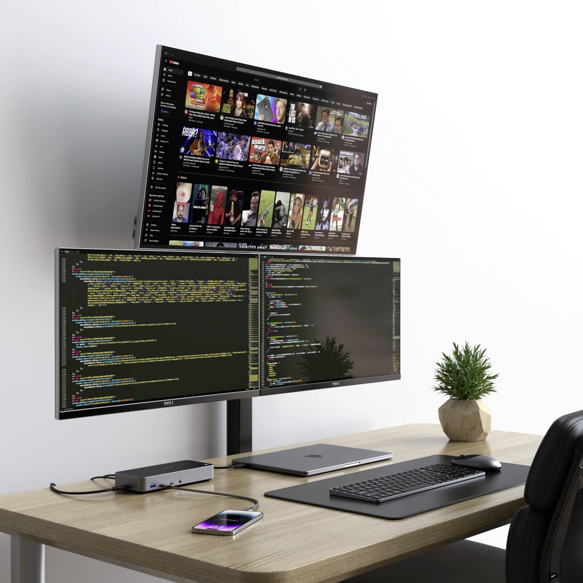 Trois écrans d'ordinateur sur un bureau : deux affichent du code de programmation, le troisième montre un portail vidéo. À côté, un ordinateur portable, un clavier, une souris et une plante.