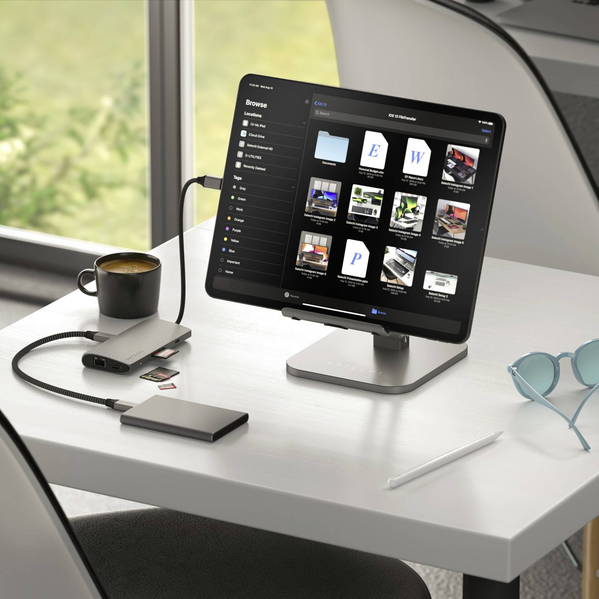 Une tablette posée sur une table avec un hub connecté et des supports de données en lecture ; à côté, des lunettes et une tasse de café.