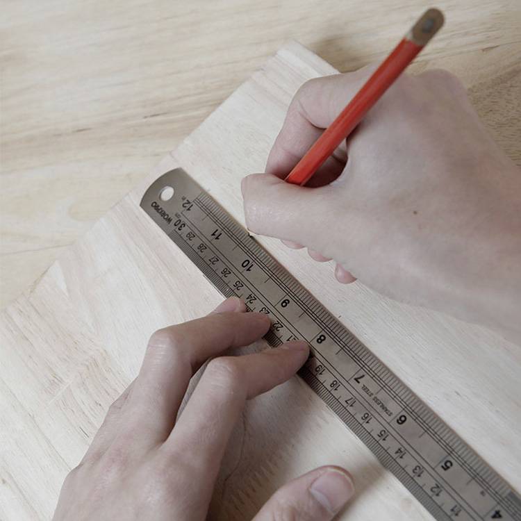 Une personne mesure avec précision des dimensions sur une planche en bois à l'aide d'un crayon rouge et d'une règle métallique.