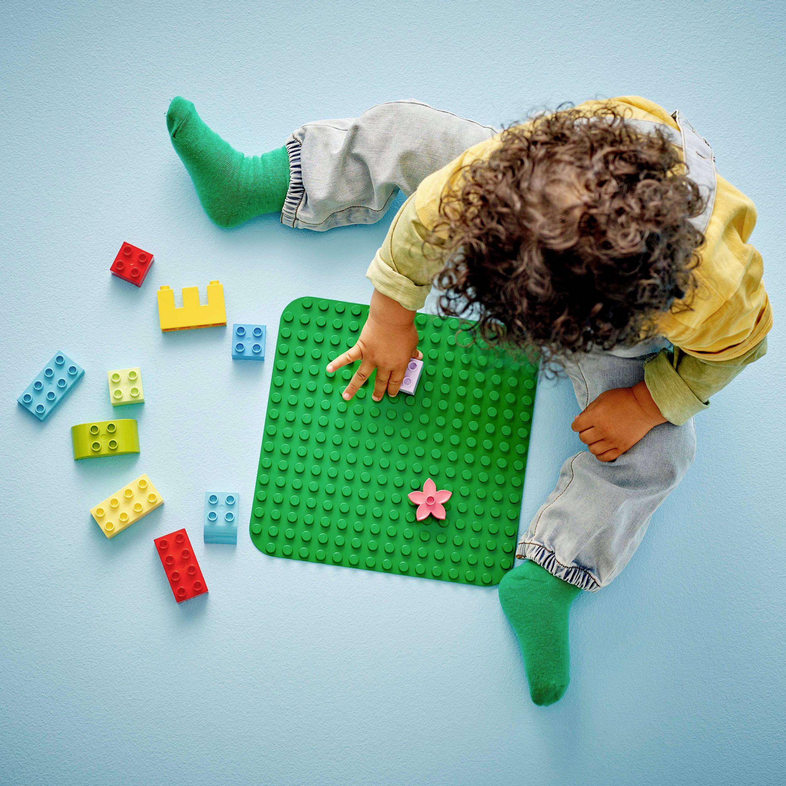 Un enfant joue sur un sol bleu avec des blocs colorés et une plaque de construction verte, sur laquelle se trouve un morceau de fleur rose.